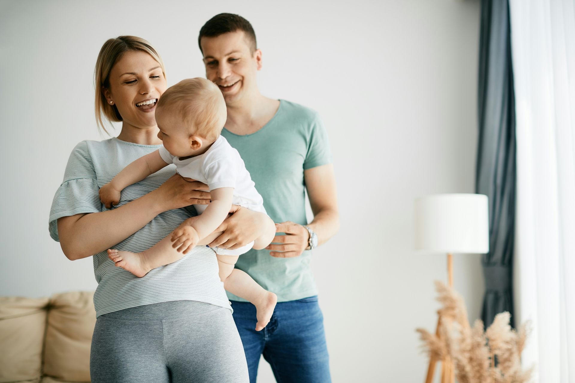 Woman holding baby; man smiles. They stand inside a room with a lamp and window.