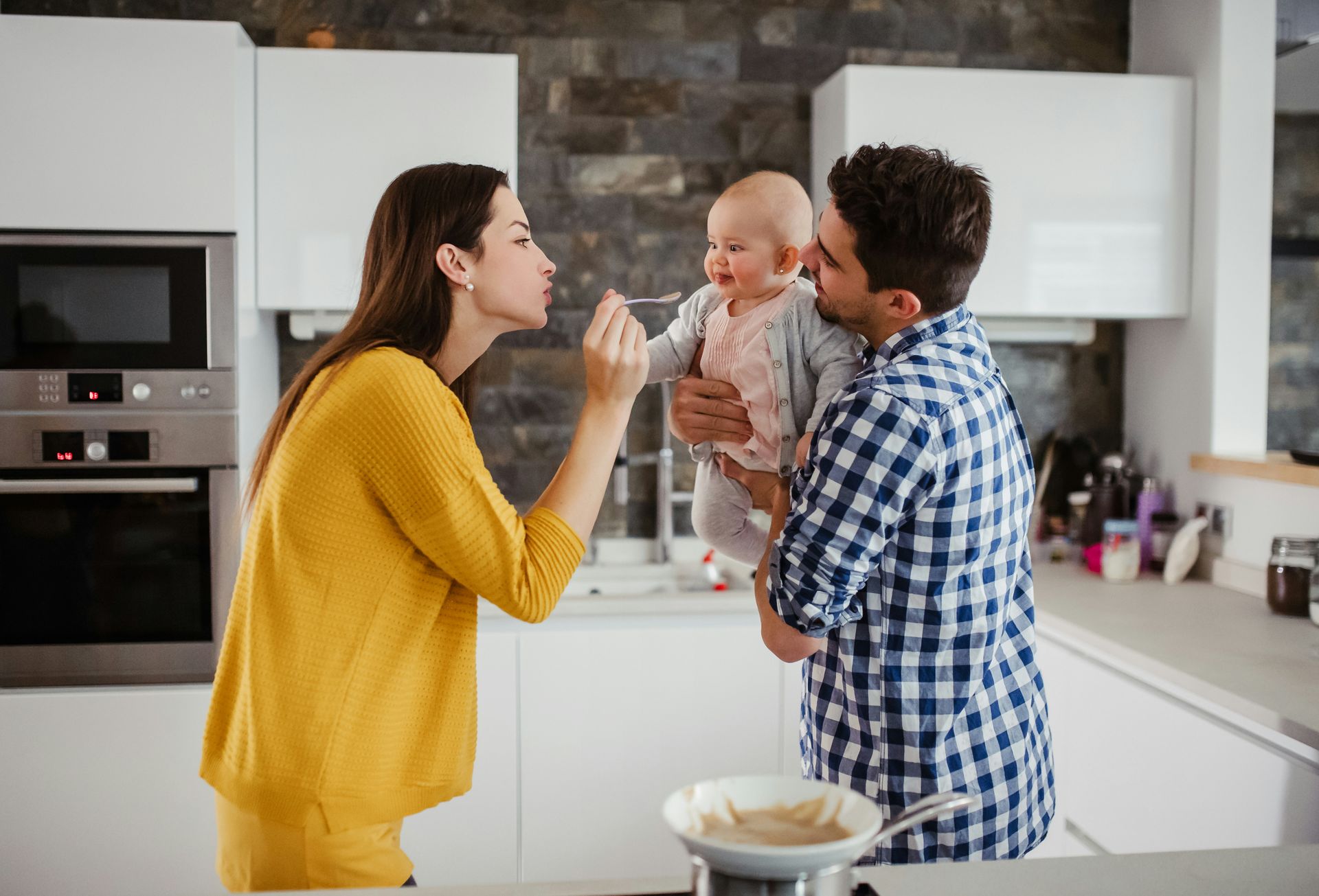 Parents in modern kitchen, holding and interacting with a baby; woman blowing kiss, man smiles.