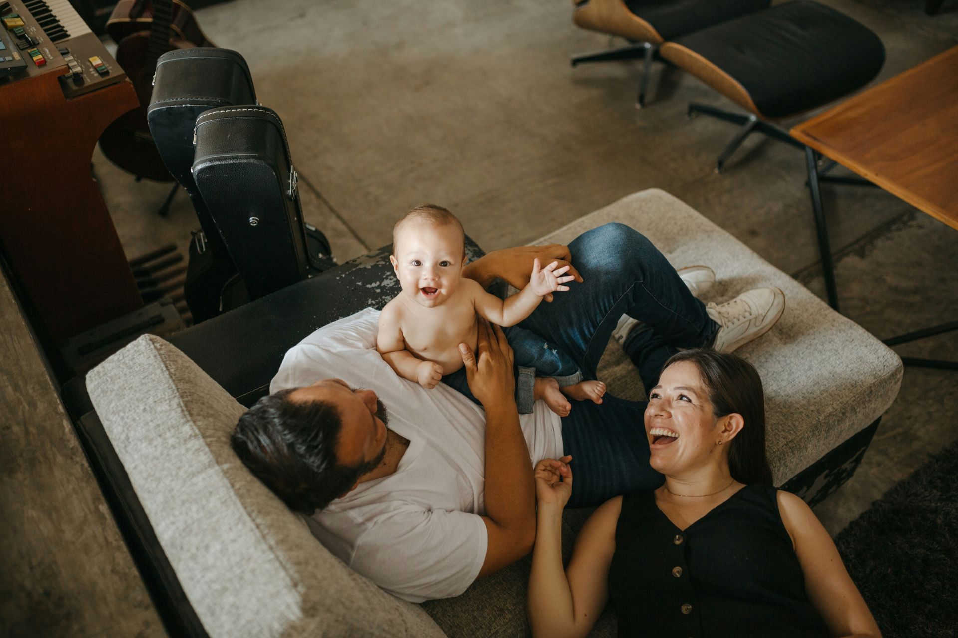 Family on a couch, baby sits on top. Parents are smiling and interacting with the baby indoors.
