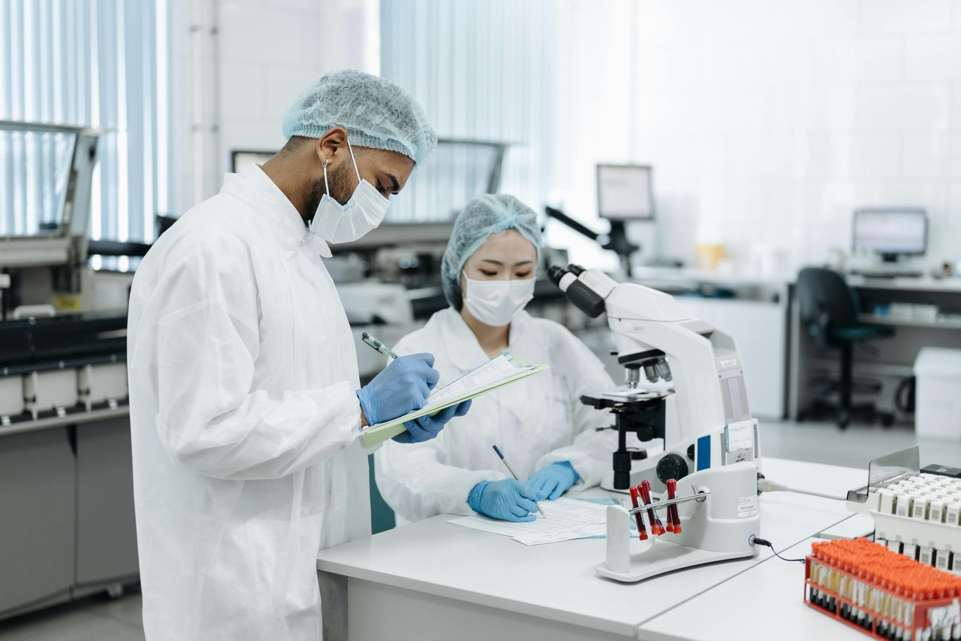Two lab workers in white coats and masks examining samples, writing notes in a lab setting.
