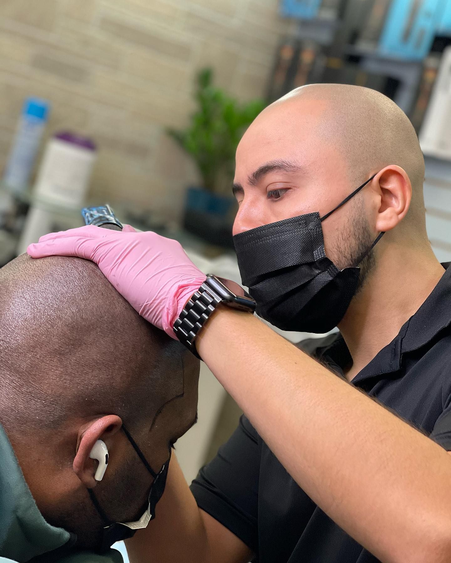 A man wearing a mask and pink gloves is getting a tattoo on his head.