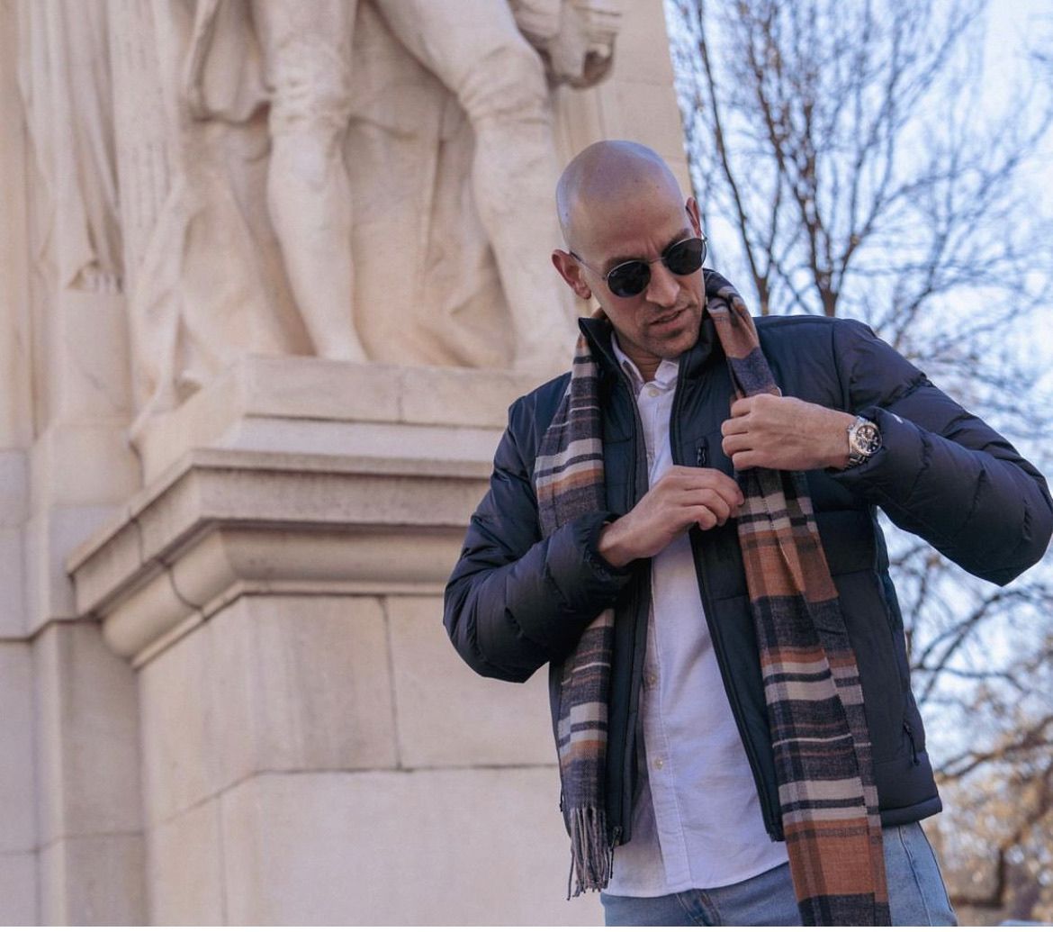 A man wearing sunglasses and a scarf is standing in front of a statue.