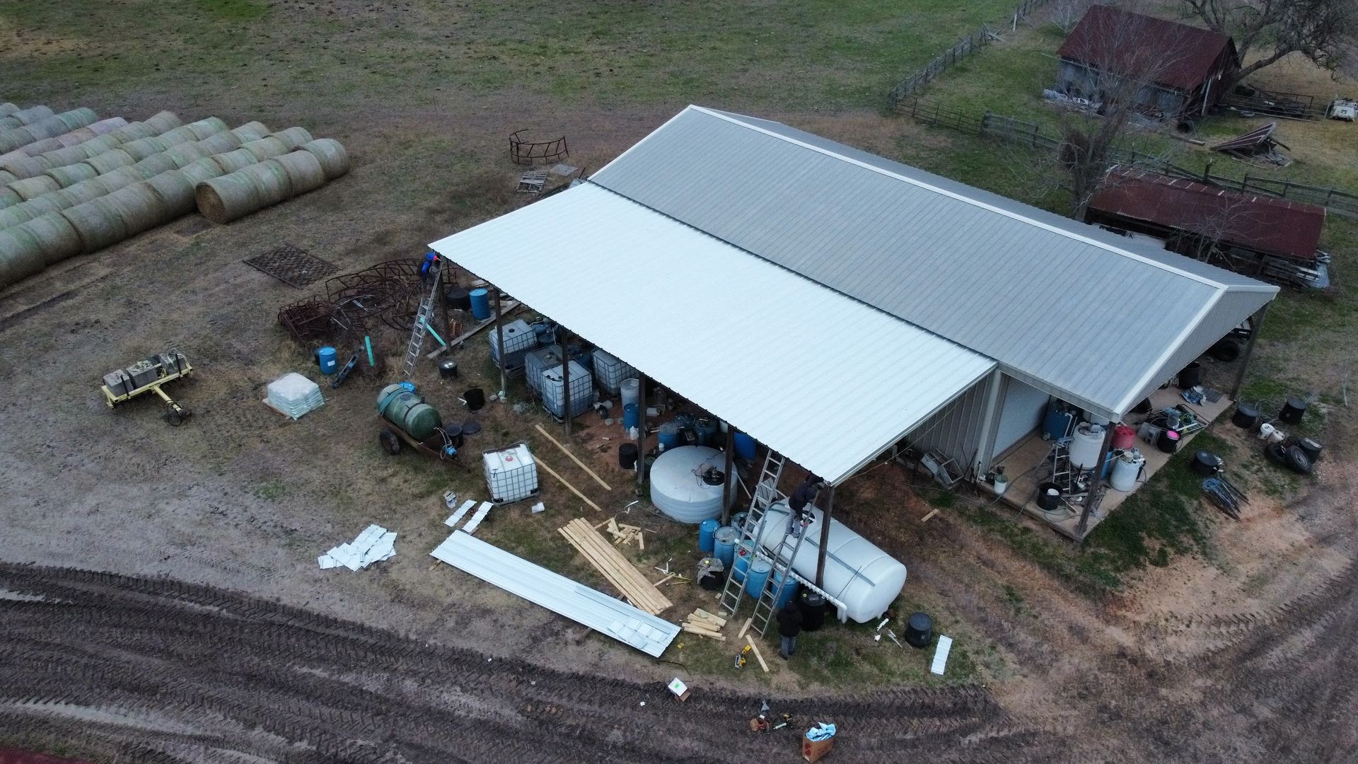 An aerial view of a farm building with a metal roof, hay bales, and farming equipment in a rural setting.