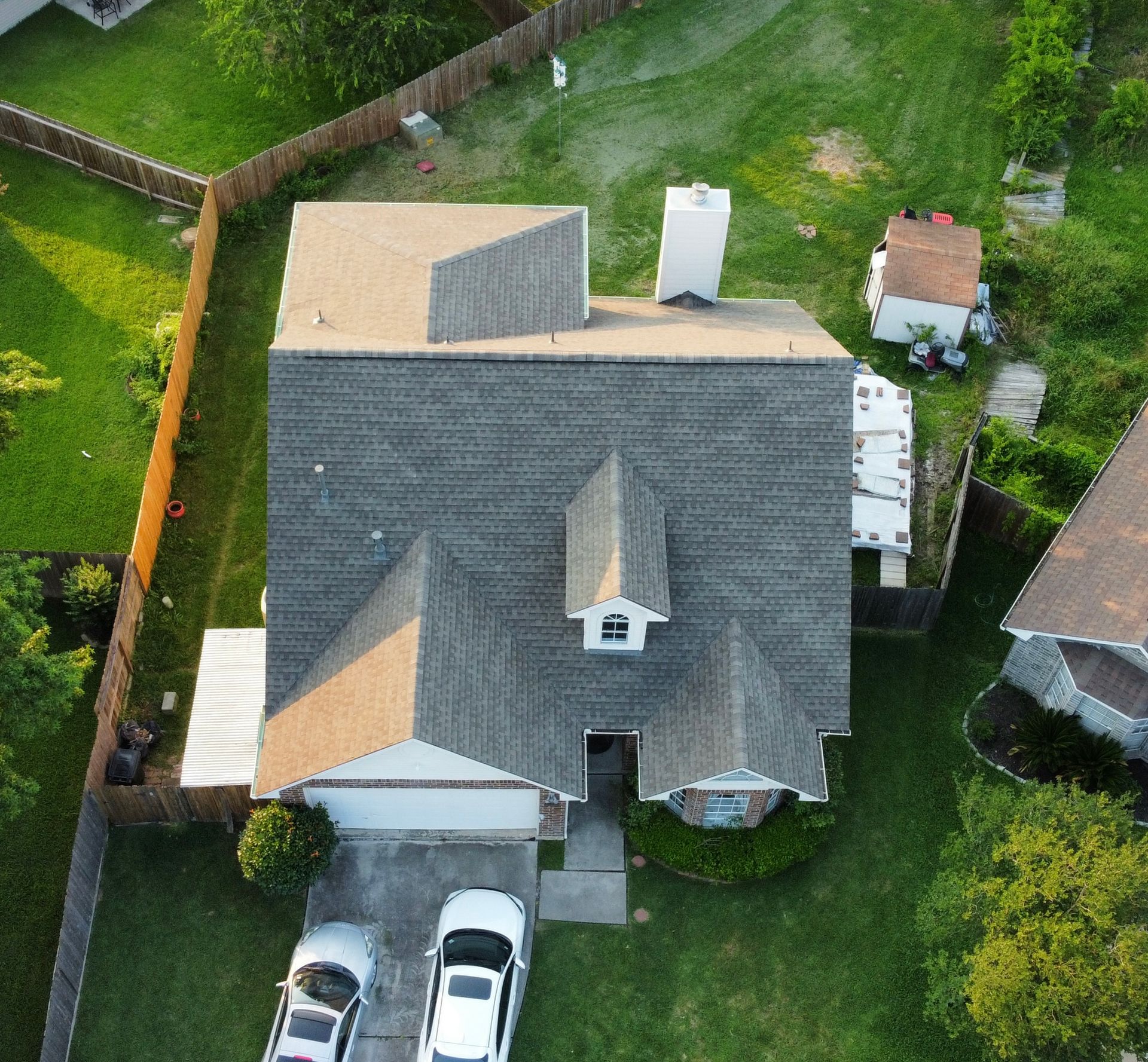 Aerial view of a house with a gray roof, white chimney, and two cars parked in the driveway. Green grass surrounds the house.