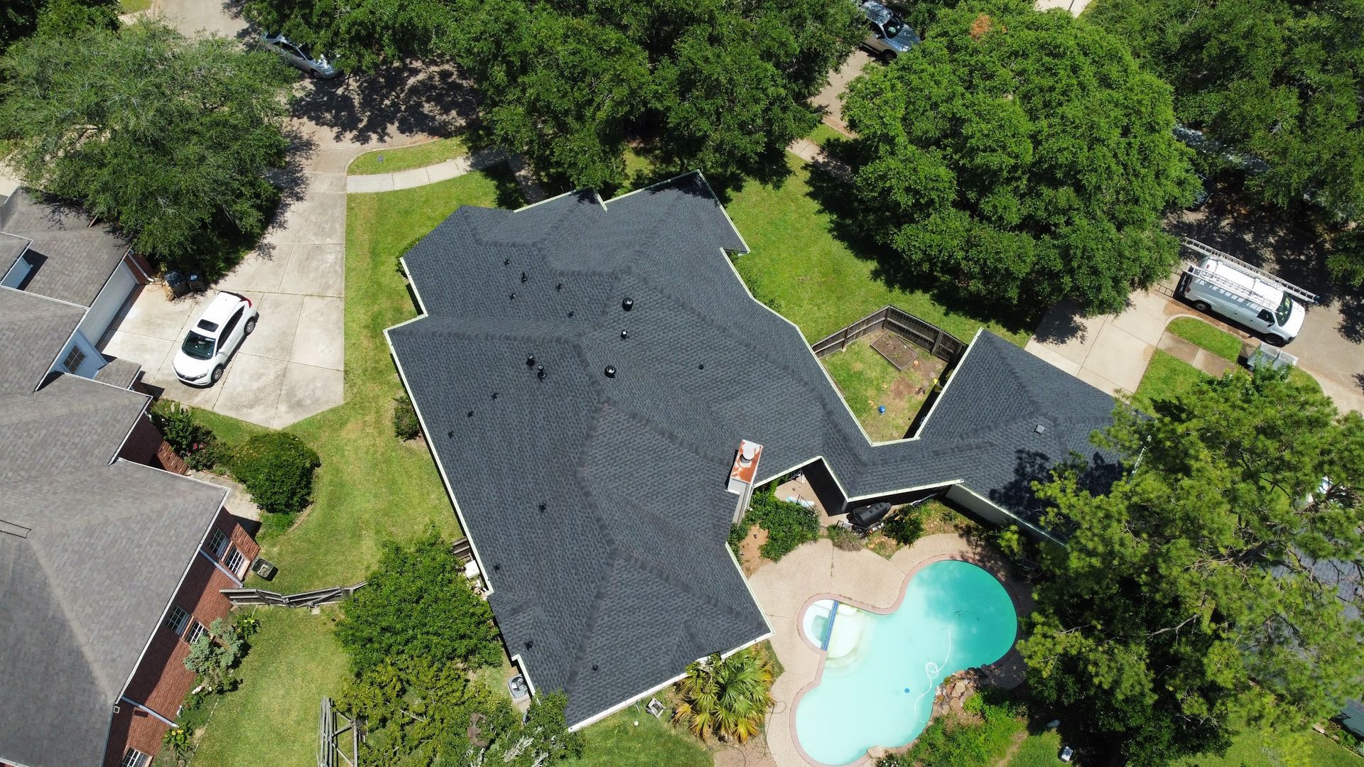 Aerial view of a house with a dark roof and swimming pool surrounded by green lawn and trees.