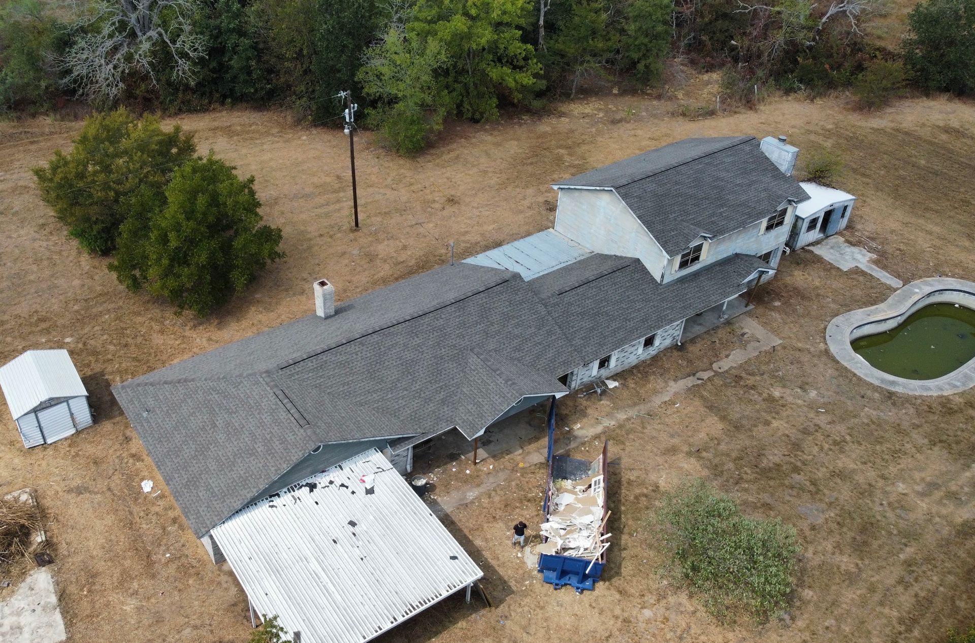 Aerial view of a two-story house with a gray roof, a small pool, and dry, brown grass surrounding it. A shed is to the left.