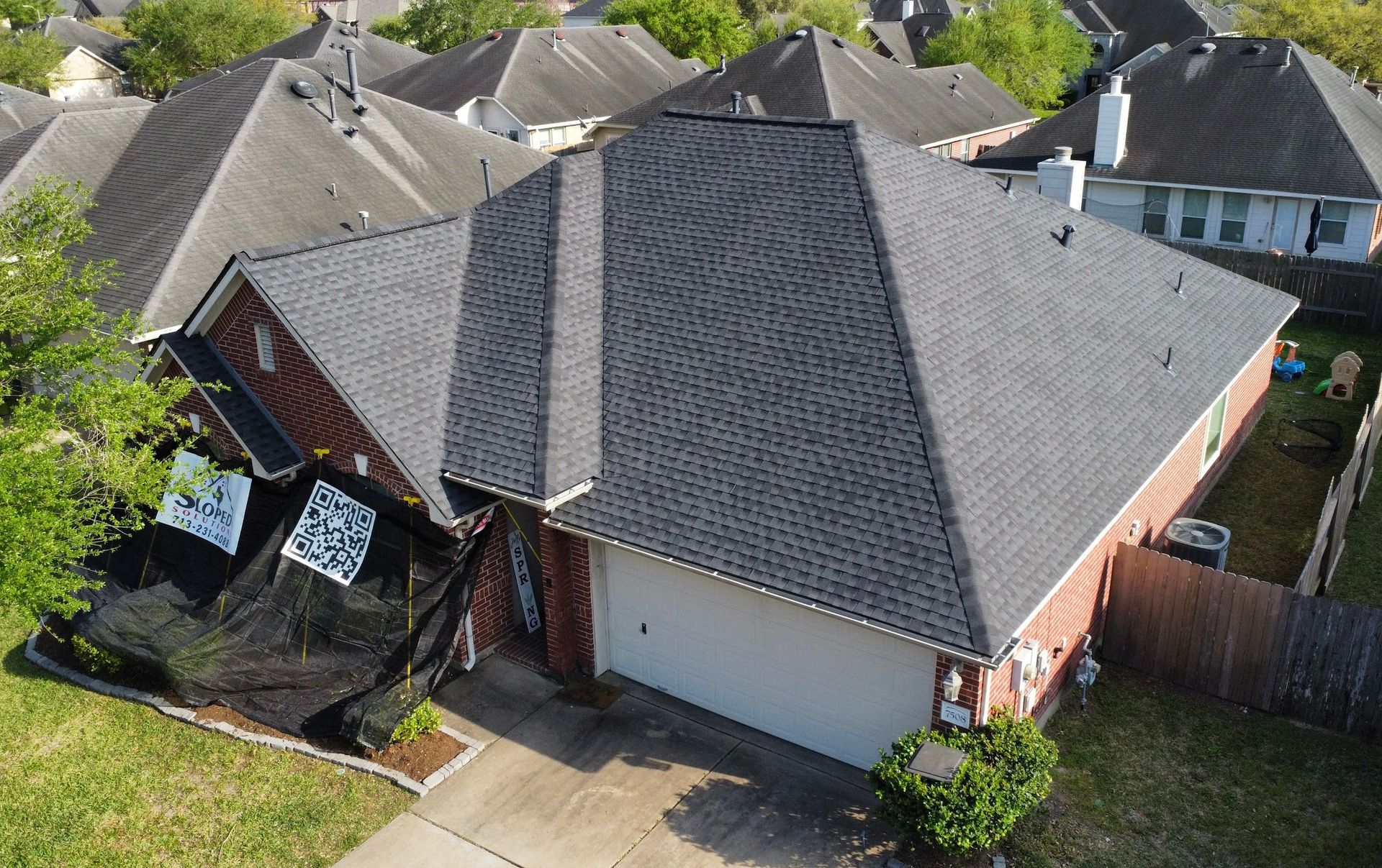 Aerial view of a brick house with a dark gray shingle roof. Black netting covers part of the side.
