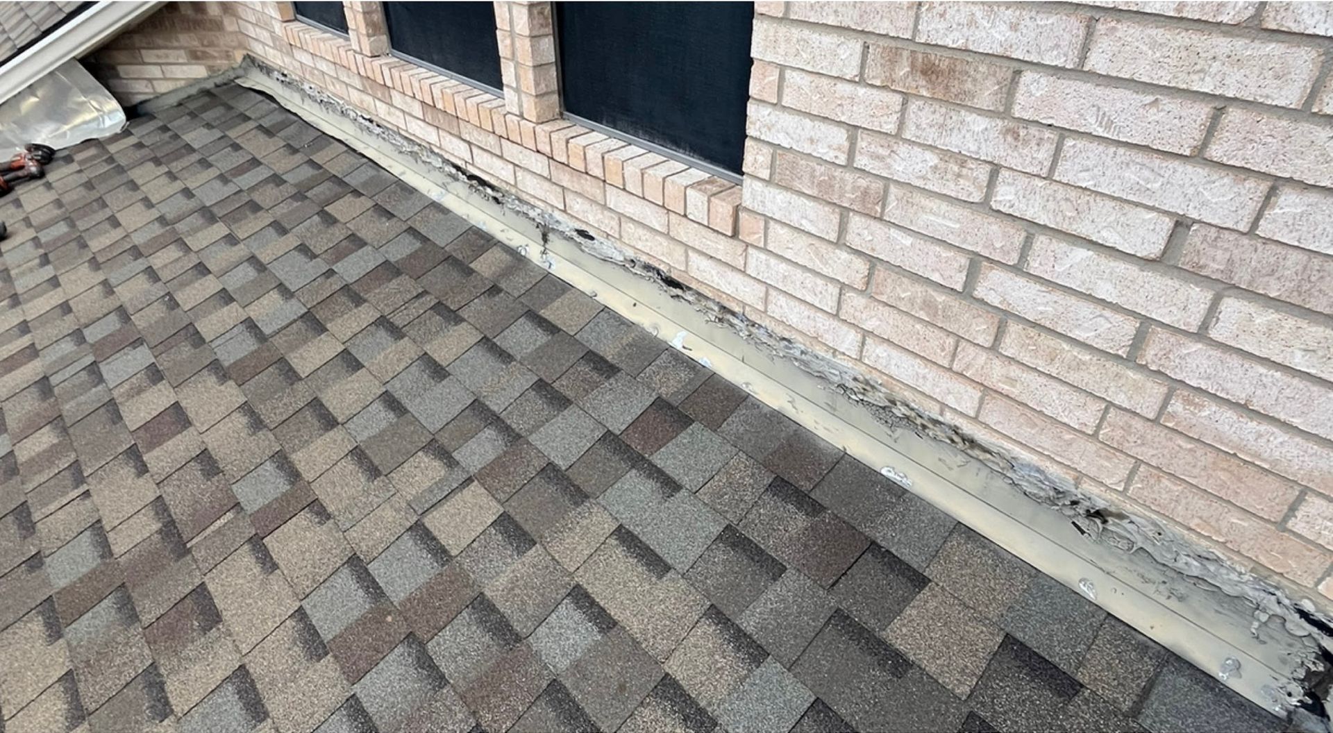 Roof shingles meet brick wall, with a strip of sealant visible along the joint.