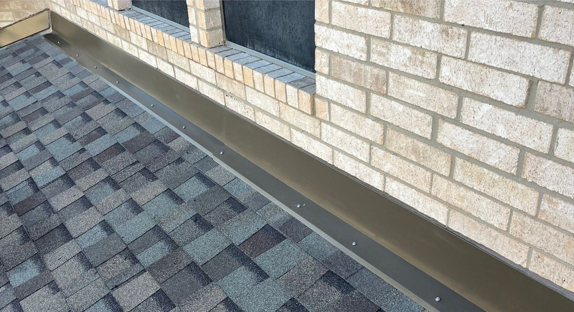 Close-up of a roof with shingles and a dark-colored metal flashing against a brick wall.