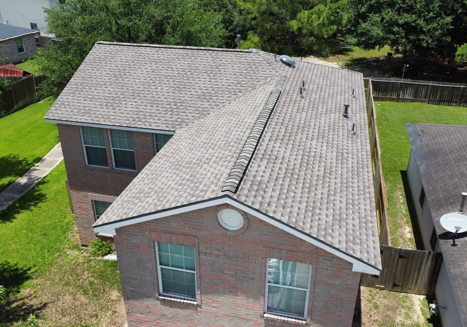 Aerial view of a two-story brick house with a gray shingled roof. It sits on a green lawn, surrounded by trees and a fence.