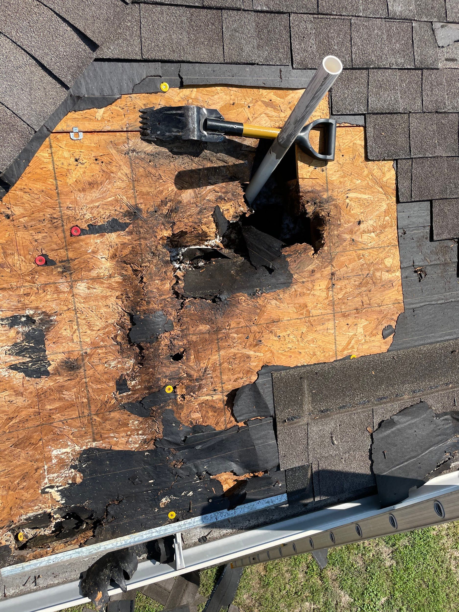 Damaged roof section with exposed wood. Shingles removed, revealing a hole, and a tool on top.