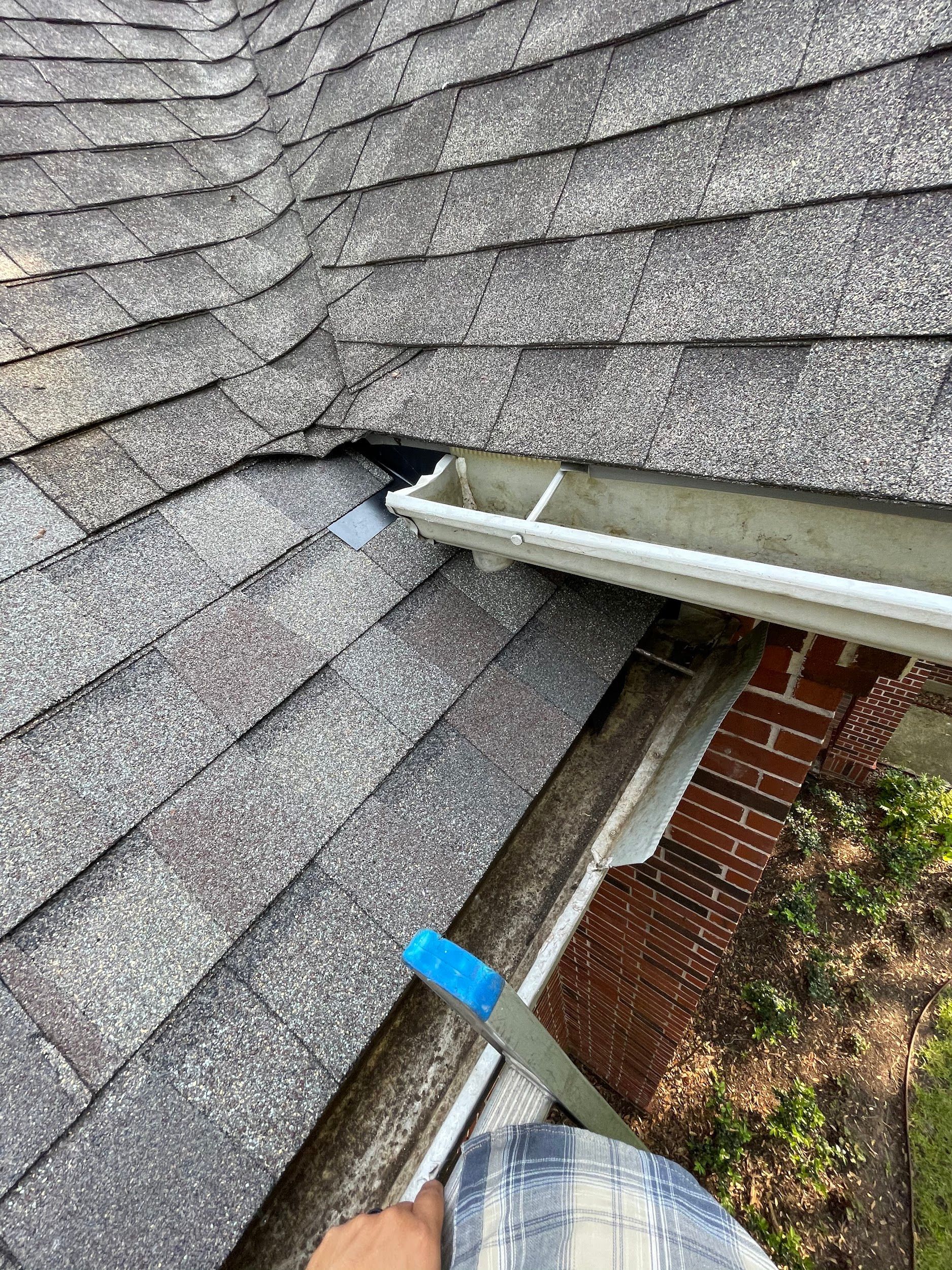 Close-up view of a roof with shingles and a gutter. Someone is in the gutter, holding a blue tool.
