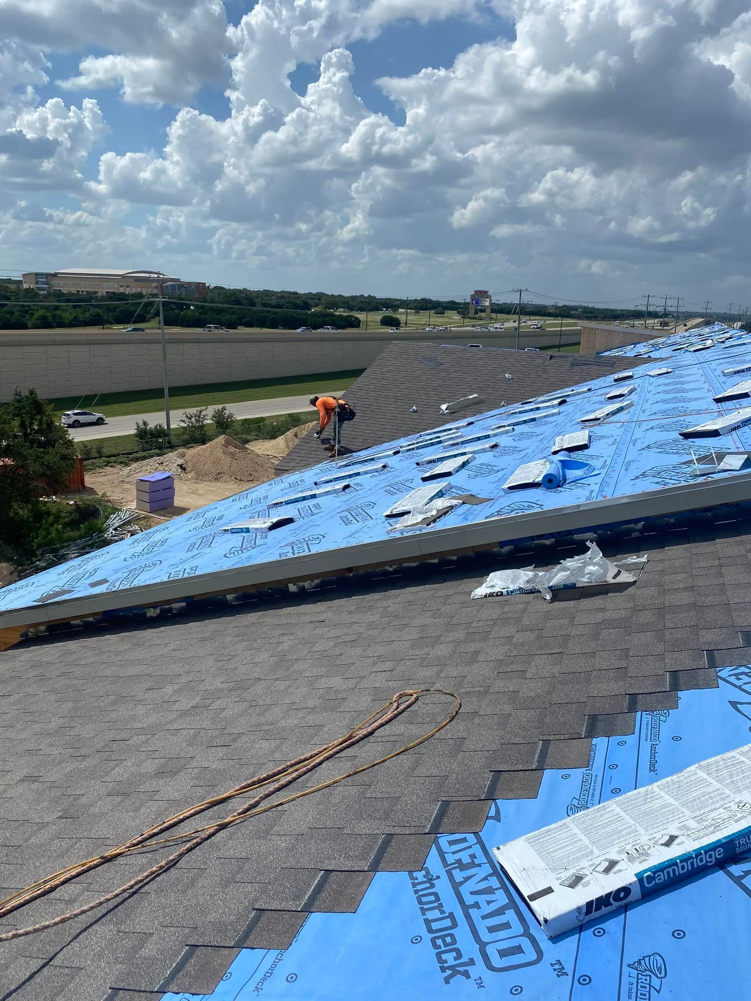Roofer working on a house roof, installing shingles over blue underlayment, on a sunny day. 