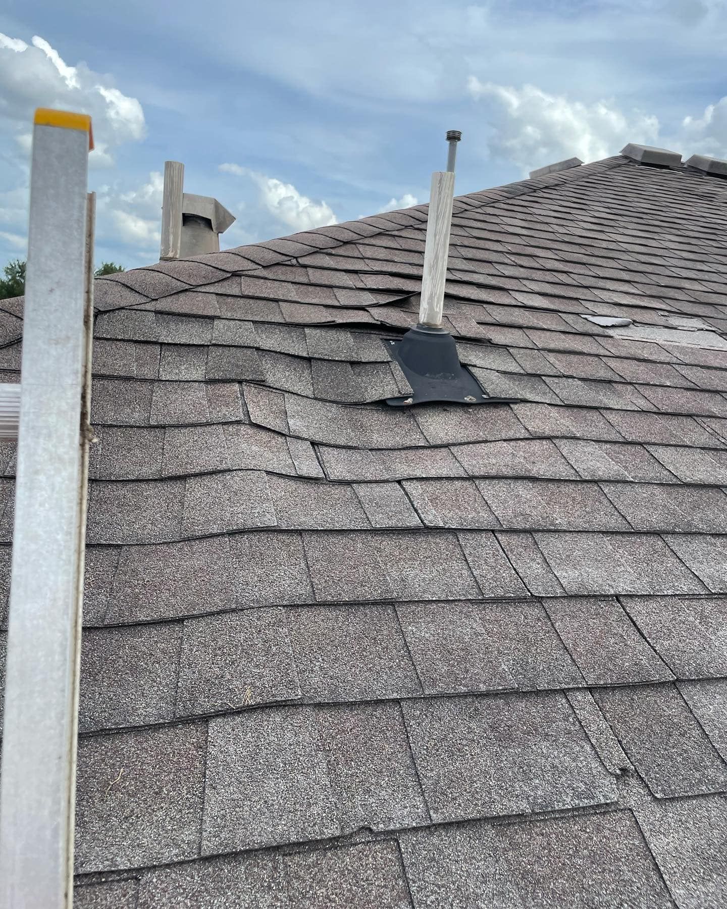 Damaged asphalt shingle roof with a black patch around a pipe. A ladder leans against the roof.