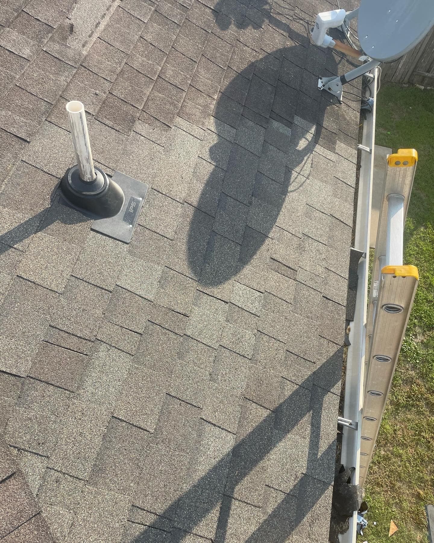 A roof with a satellite dish, plumbing vent, and ladder; casting shadows on the aged, grey asphalt shingles.