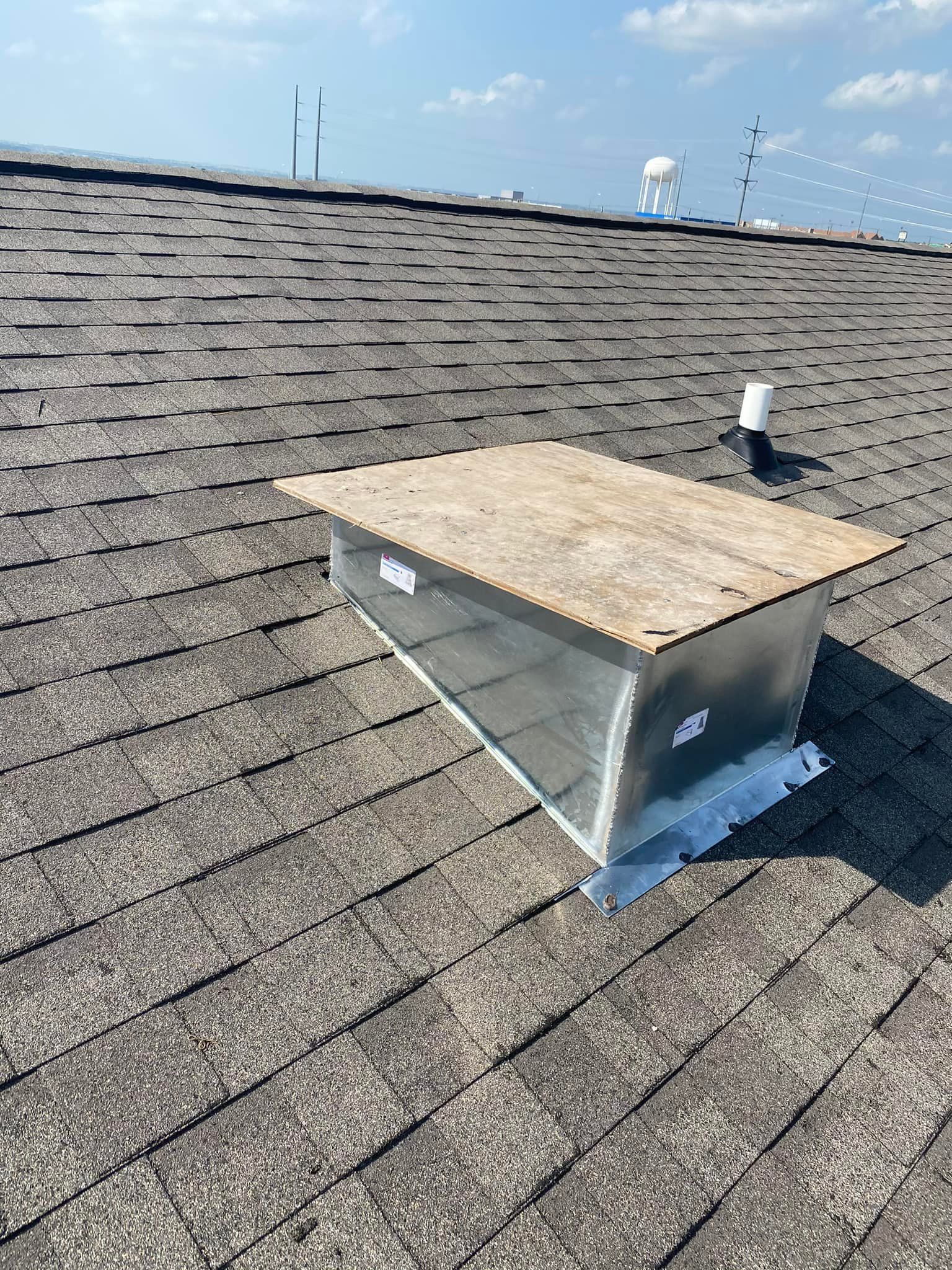 A metal vent box on a brown shingle roof. The sky is blue with wispy clouds.