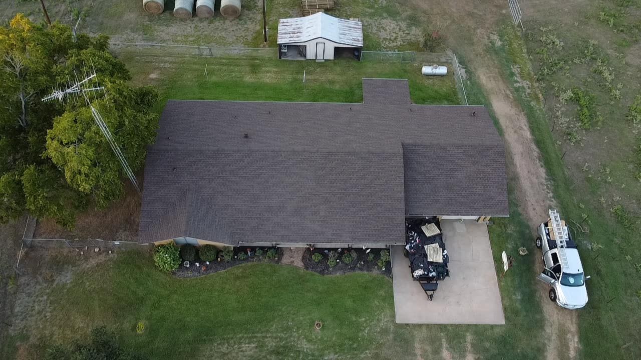 Aerial view of a house with a brown roof, a driveway with two trucks, and surrounding green grass.