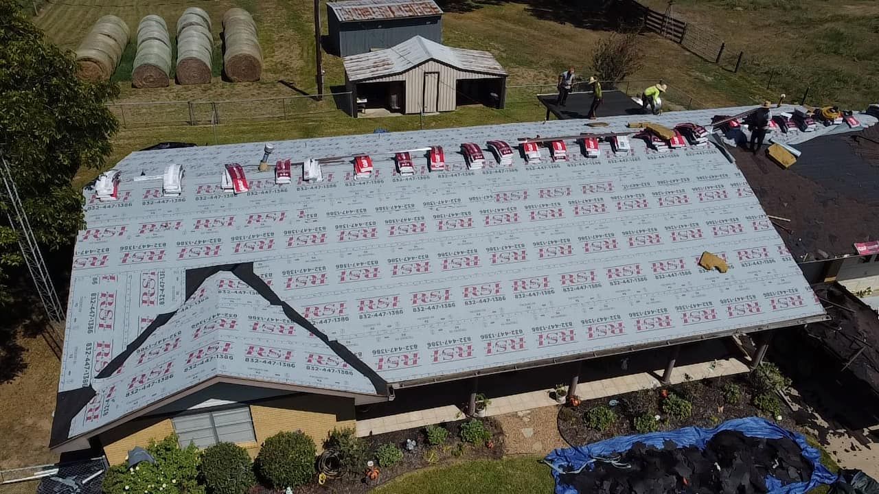 Aerial view of a building with workers replacing a roof. Rows of gray roofing material are visible, as well as red markers.