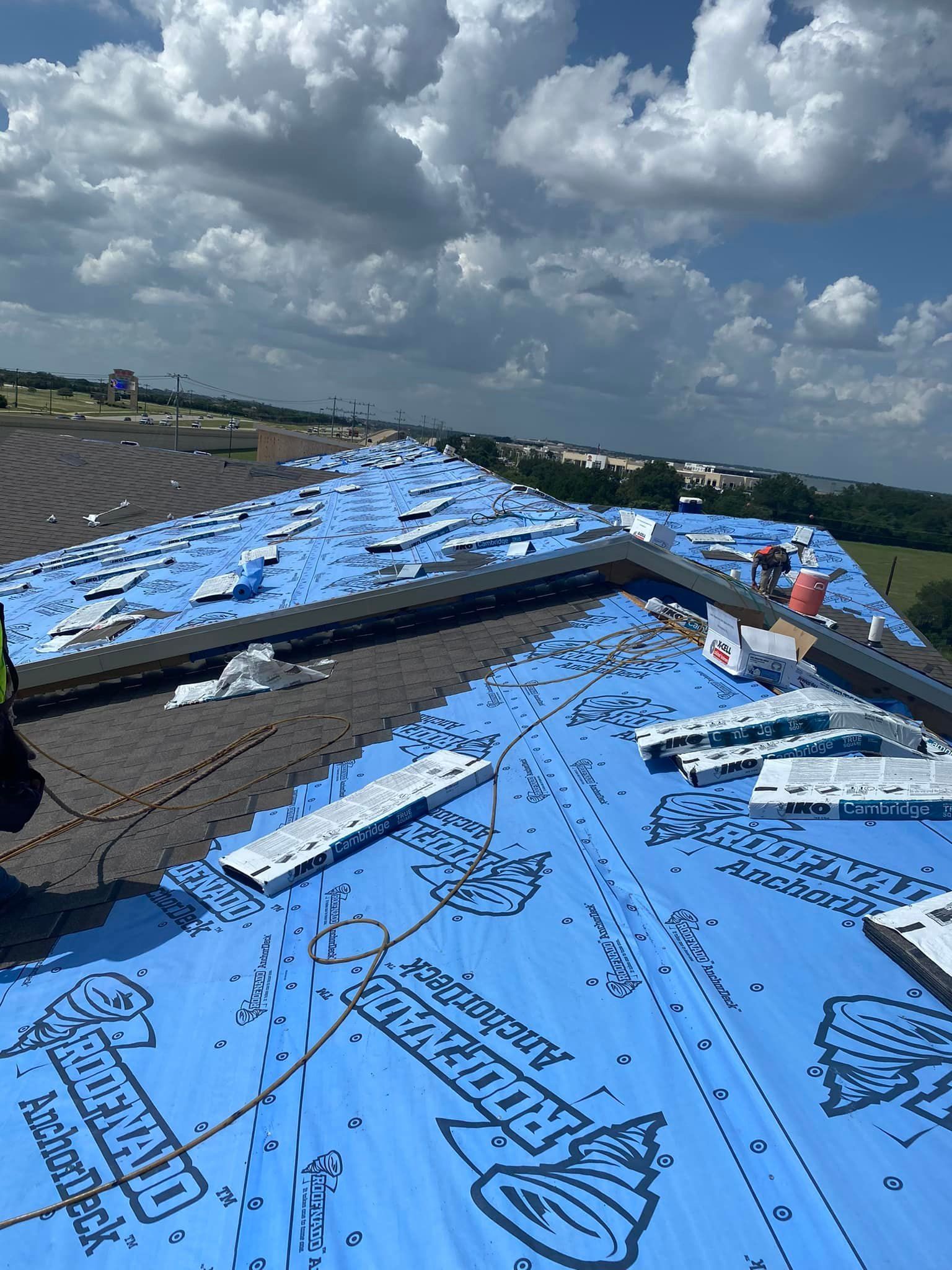 Roofing work in progress on a rooftop under a cloudy sky. Blue underlayment covers the roof.