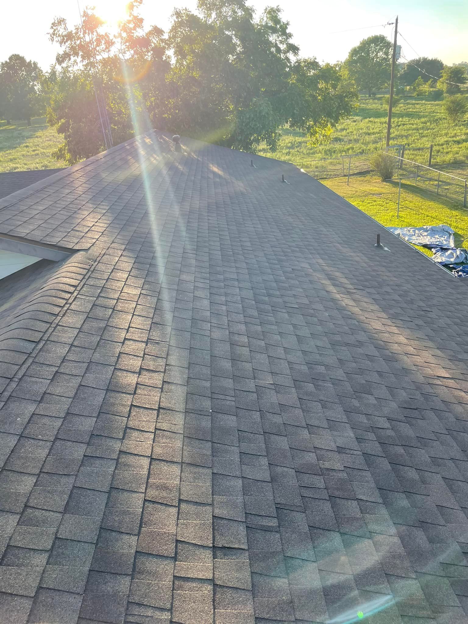 Sunlit view of a dark gray shingle roof, with the sun shining brightly in the background over trees and grass.