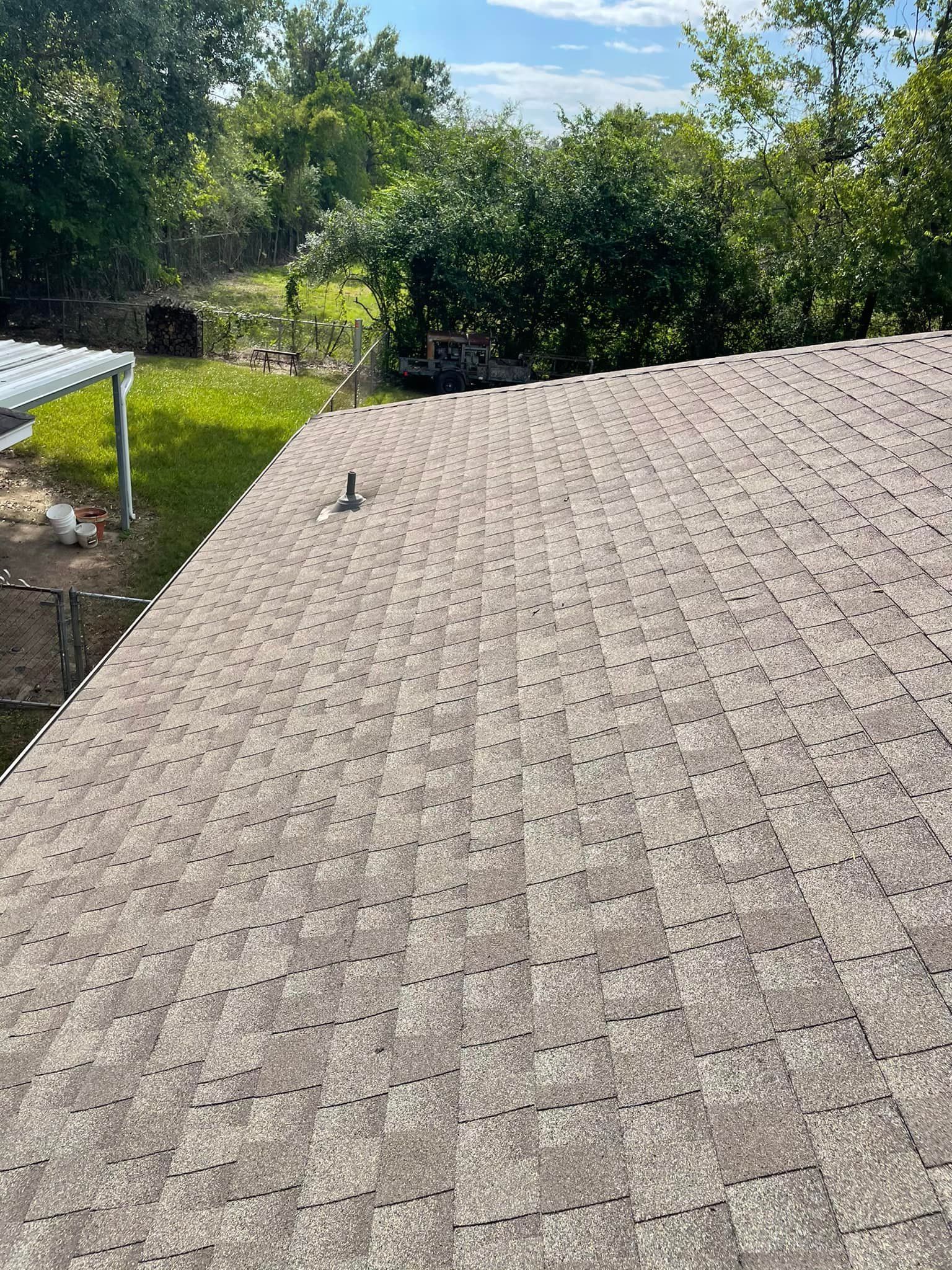 Overhead view of a weathered, gray shingle roof with a vent pipe. Green trees and grass are visible in the background.