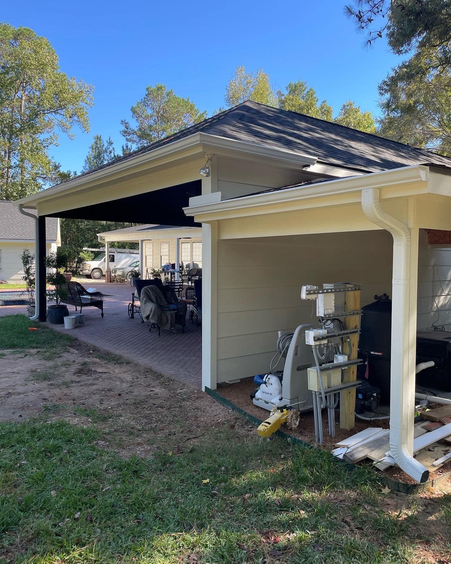 Backyard with a shaded patio, featuring a beige building with equipment, and a paved area with furniture under a awning. 