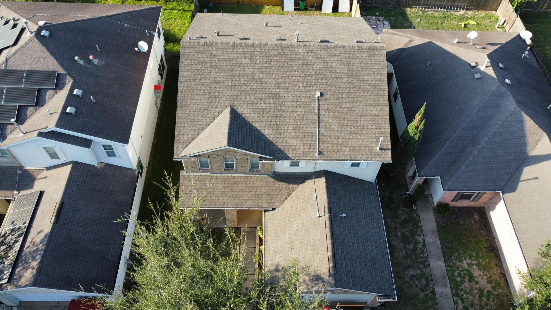 Aerial view of a two-story house with a gray roof, surrounded by other houses. Sunlight and shadows are visible.