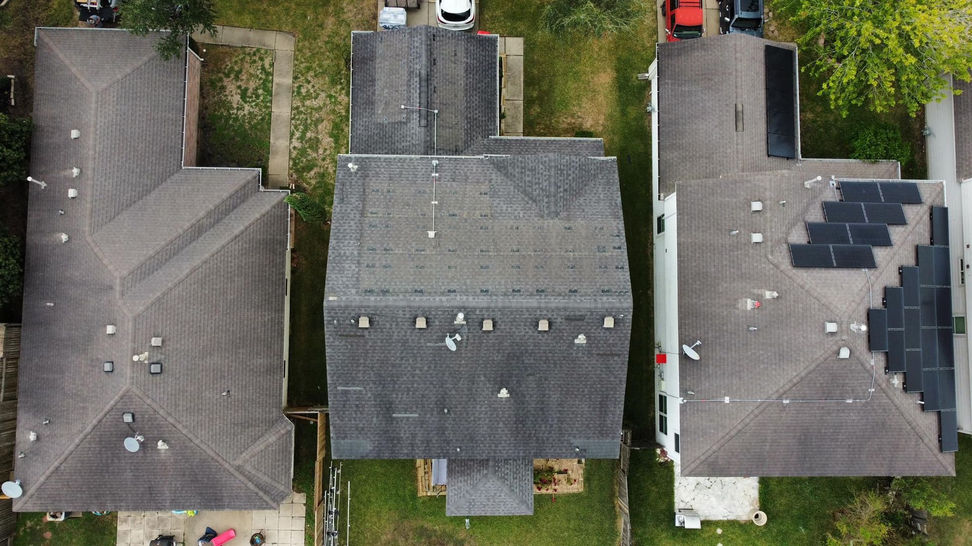Overhead view of three houses with gray roofs, surrounded by green grass. One house has solar panels.