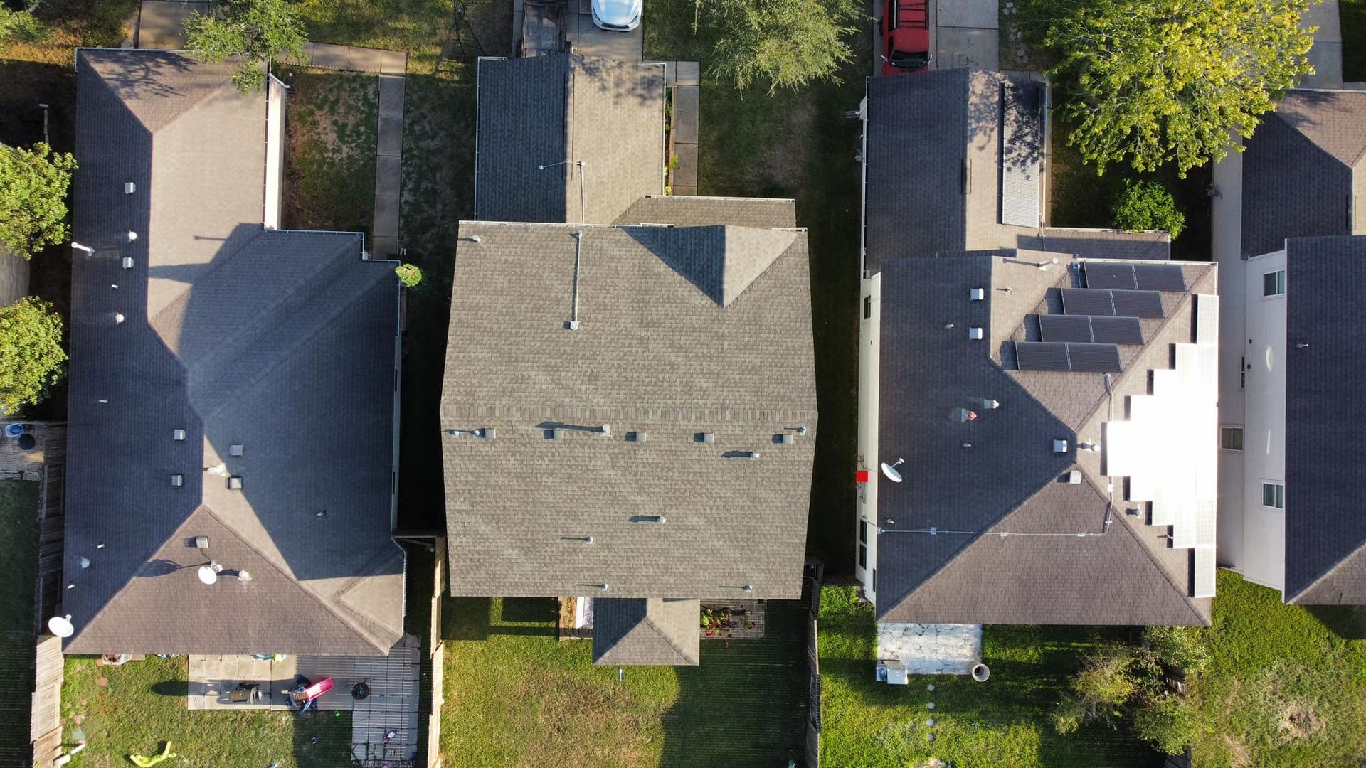 Overhead view of three houses with gray shingled roofs and small yards, located in a suburban area with some trees.