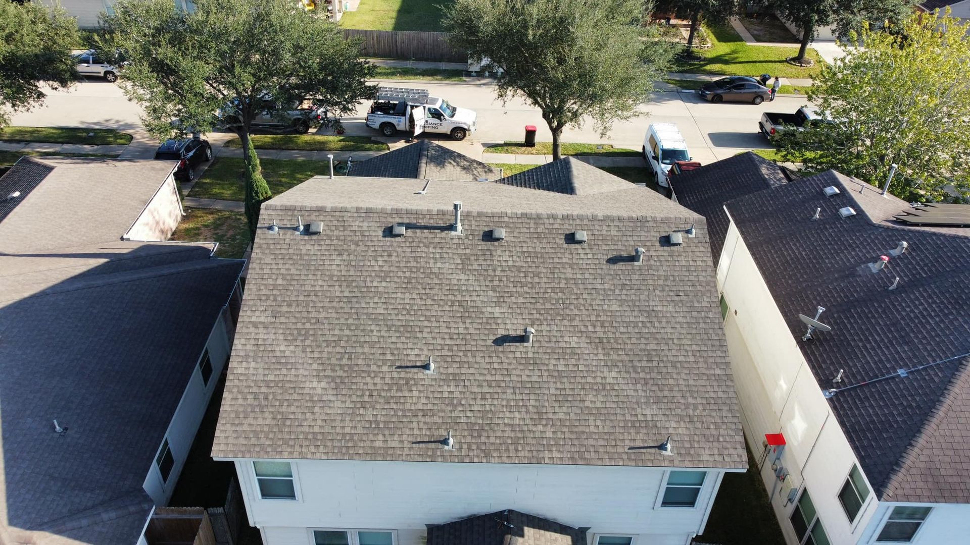 Overhead view of a two-story house with a gray shingle roof. Other houses and parked cars are visible in the background.