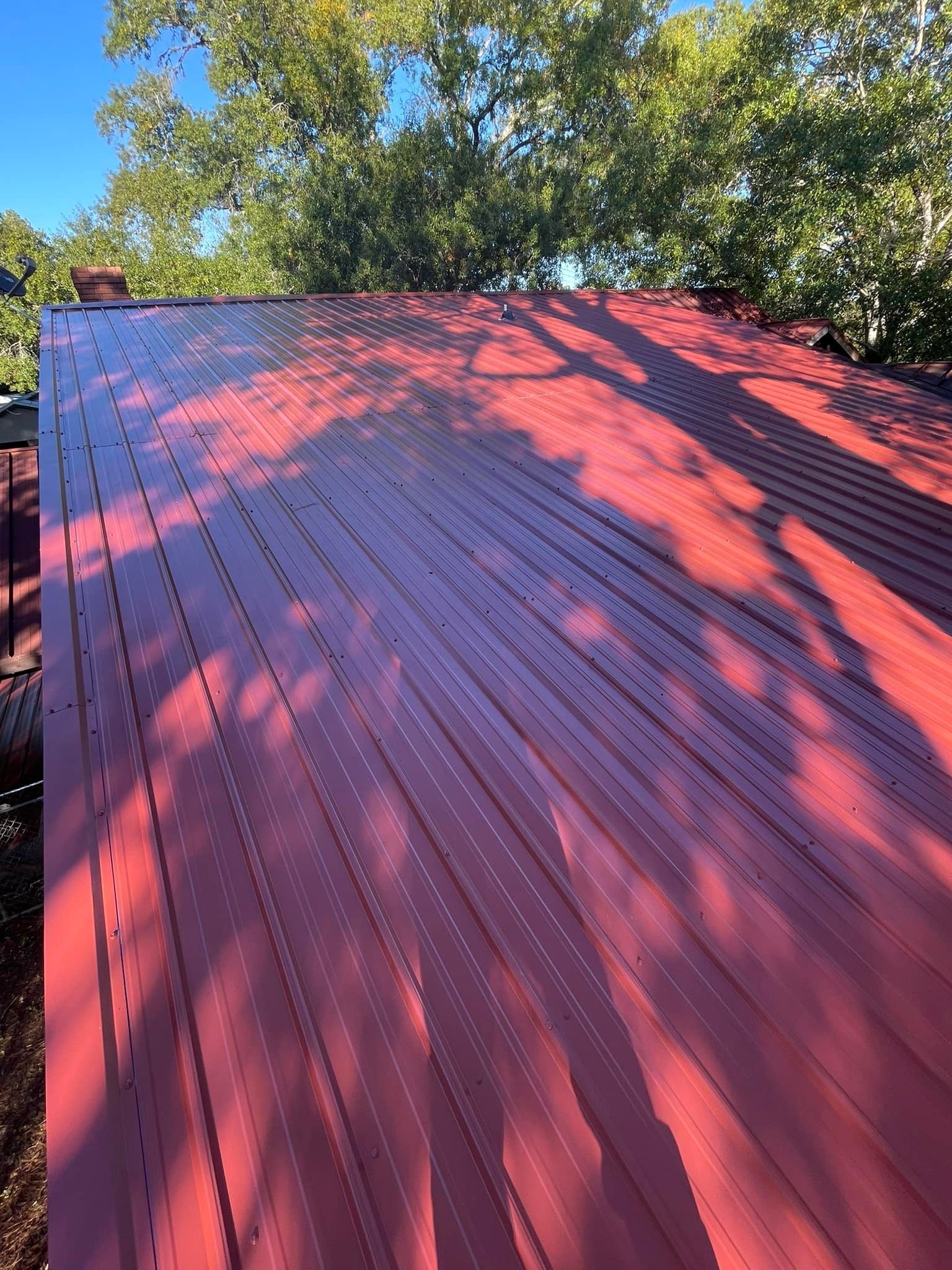 Red metal roof on a building with the shadow of trees cast across it. The sky is blue, and trees surround the roof.