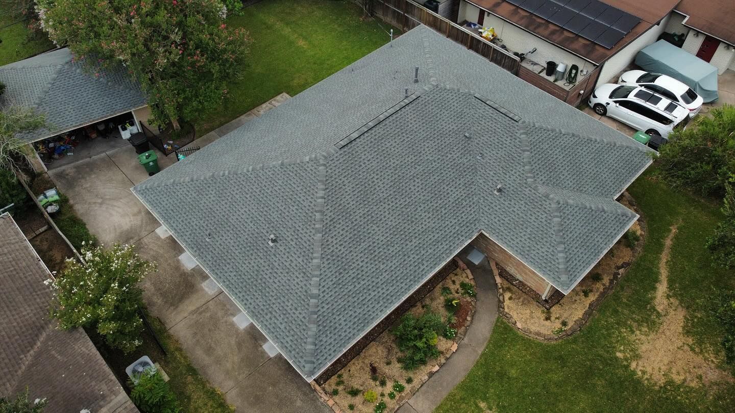 Aerial view of a house with a grey shingled roof and attached garage. Green lawn surrounds the house.