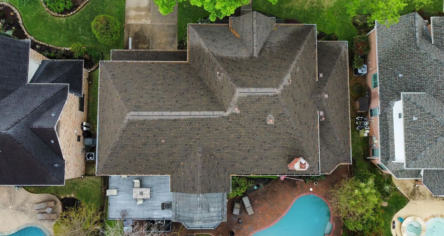 Overhead view of a large, brown-roofed house with a pool and neighboring houses. Green trees and grass surround the property.