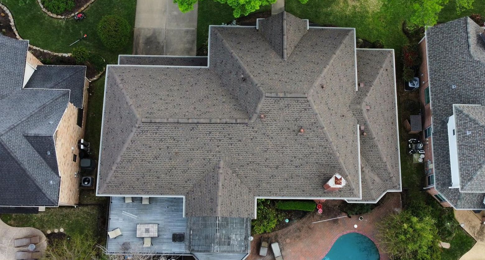 Aerial view of a residential house with a complex brown roof, surrounded by trees and other houses.
