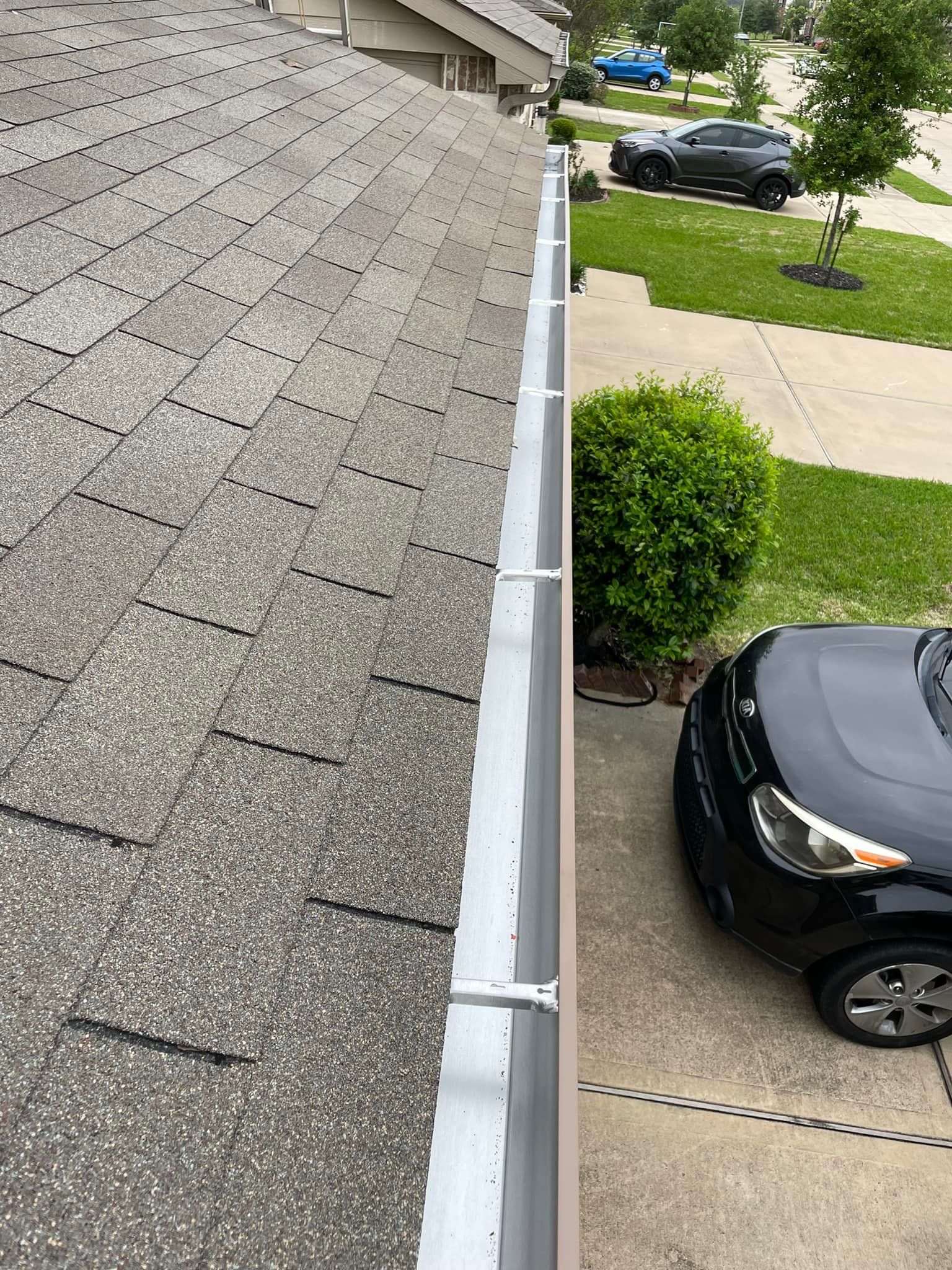 A side view of a house roof with gutters. A black car is parked on a driveway beside the gutter.