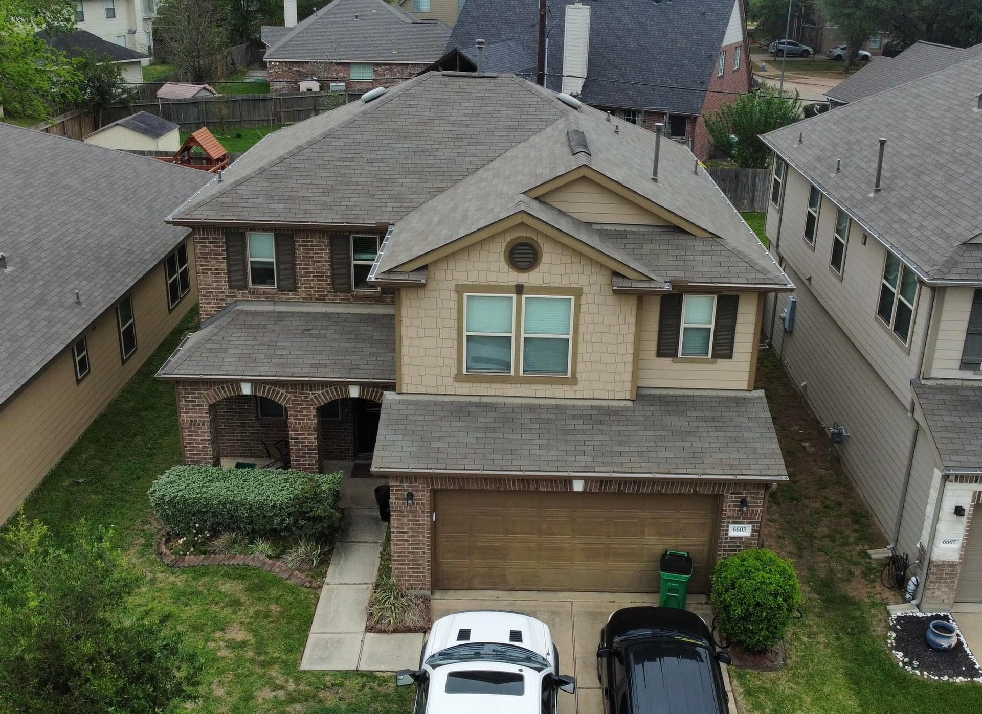 An aerial view of a two-story beige house with a brown roof and two cars parked in the driveway.