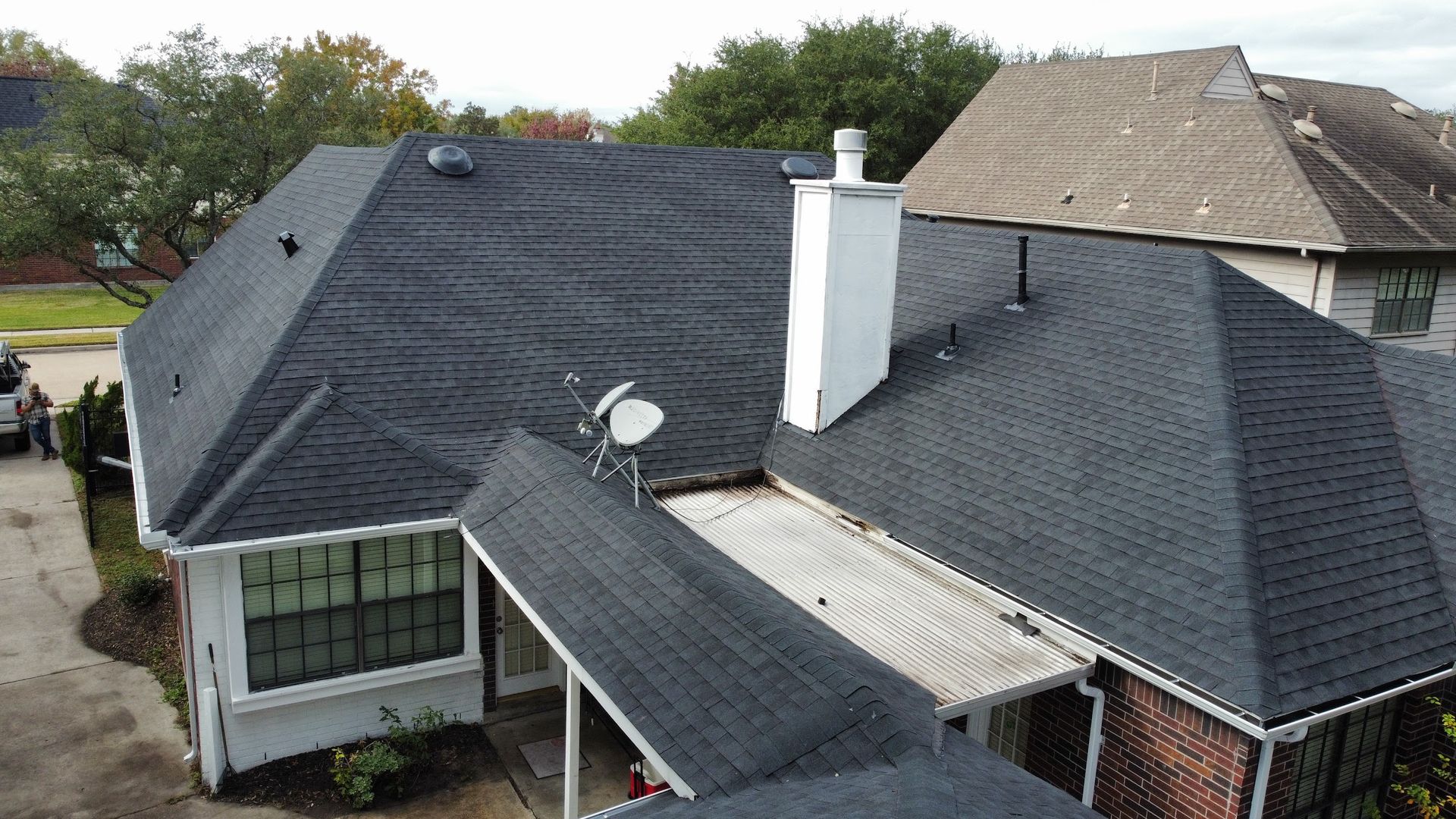 Black asphalt shingle roof on a house, with a white chimney and satellite dish. Houses and trees are in the background.