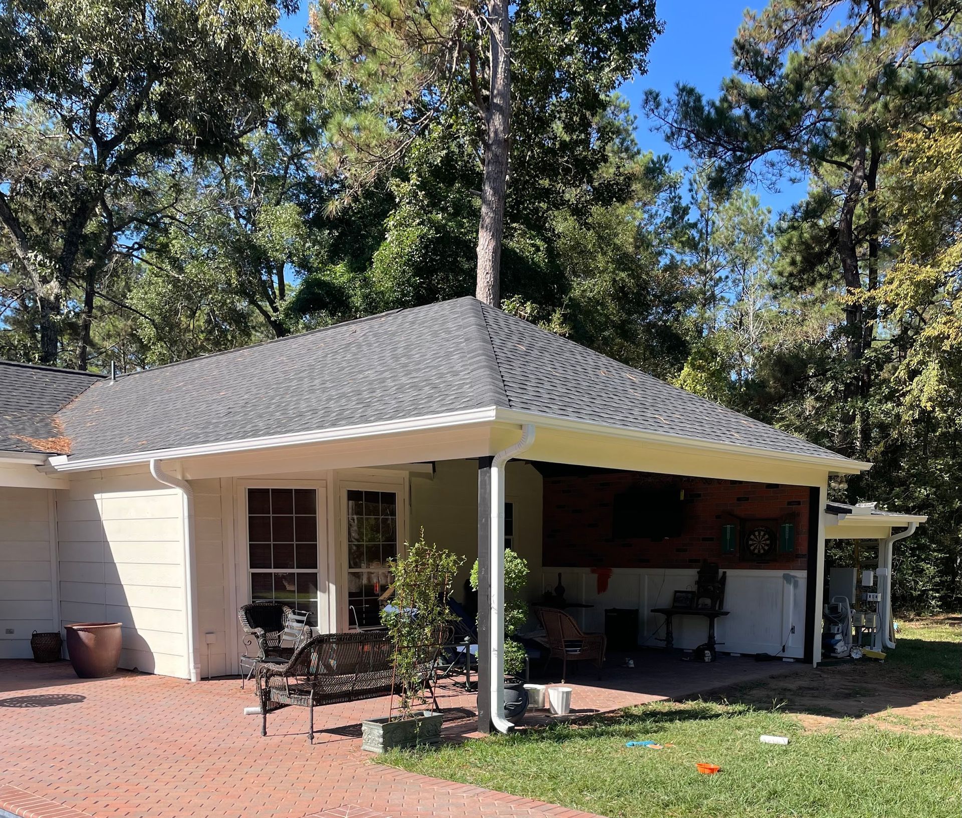 A house with a covered patio, red brick flooring, and dark gray shingles. Trees surround the house in the background.