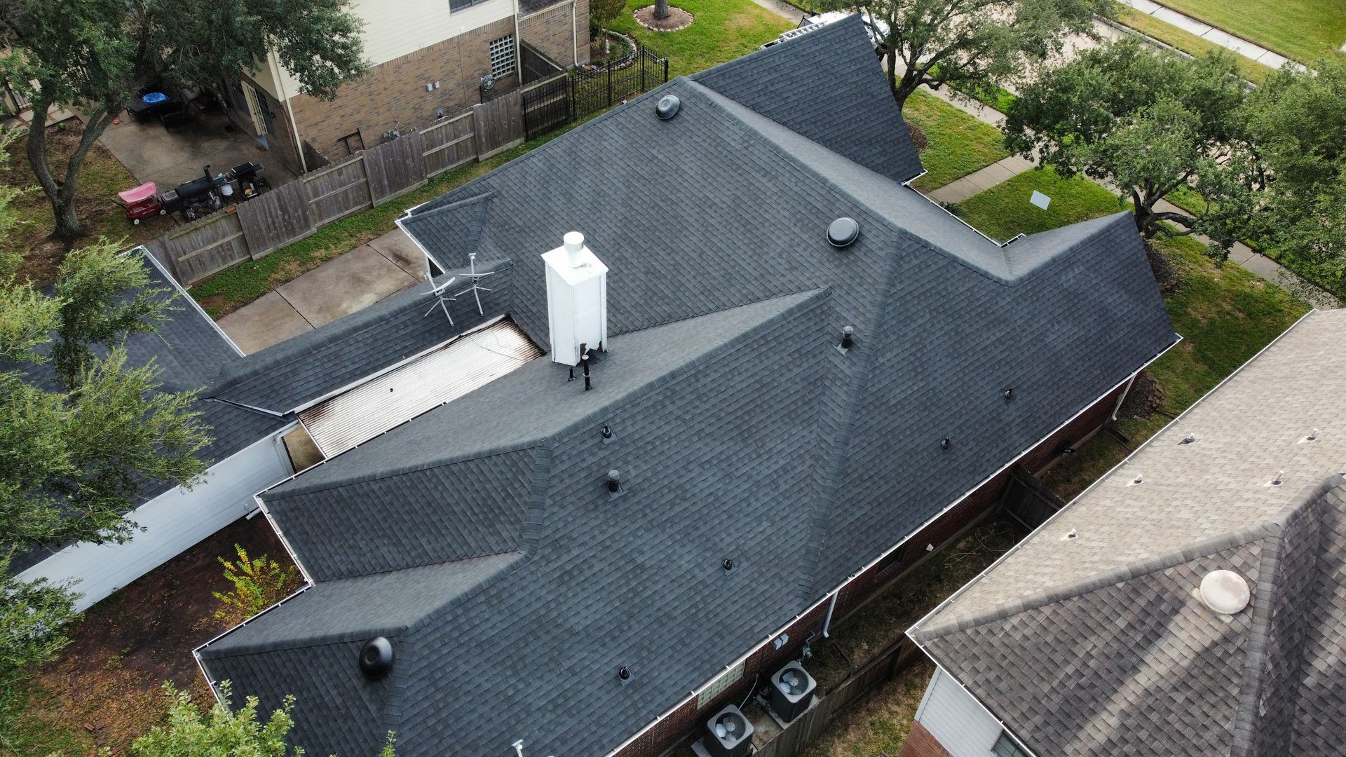 Overhead view of a dark gray asphalt shingle roof on a house with a white chimney, in a suburban neighborhood.