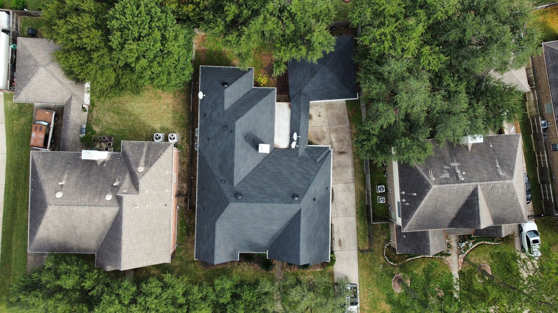 Overhead view of three houses surrounded by trees and lawns. The middle house has a dark gray roof and a paved driveway.
