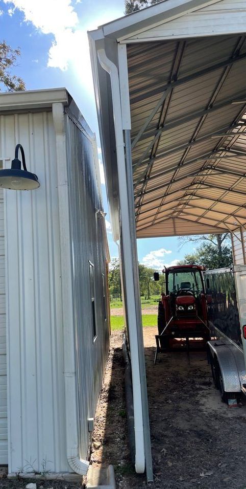 A white metal building and a metal carport are positioned close together. A red tractor is visible under the carport.