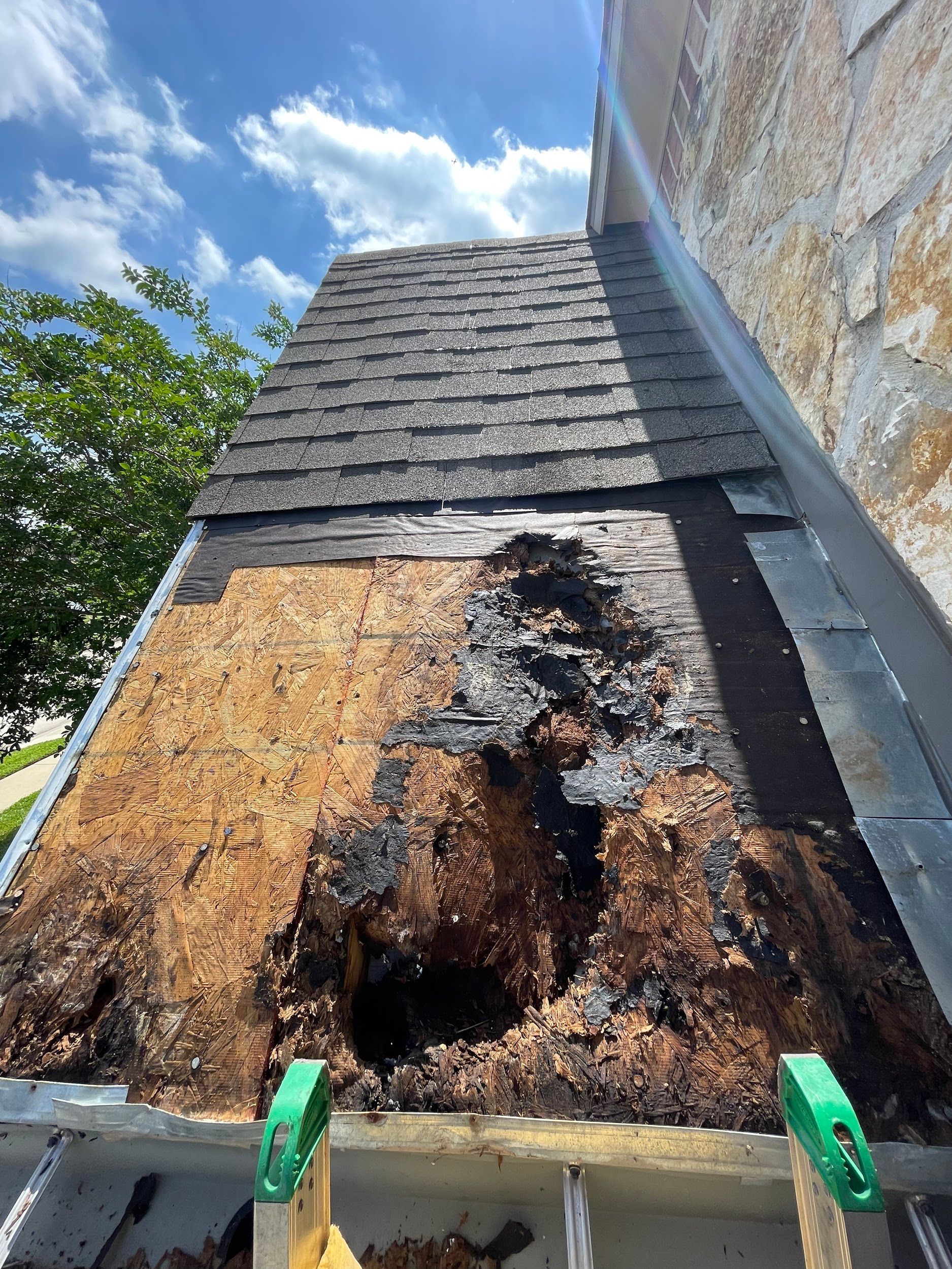 Damaged roof section with missing shingles exposing rotted wood and dark discoloration. Blue sky is in the background.