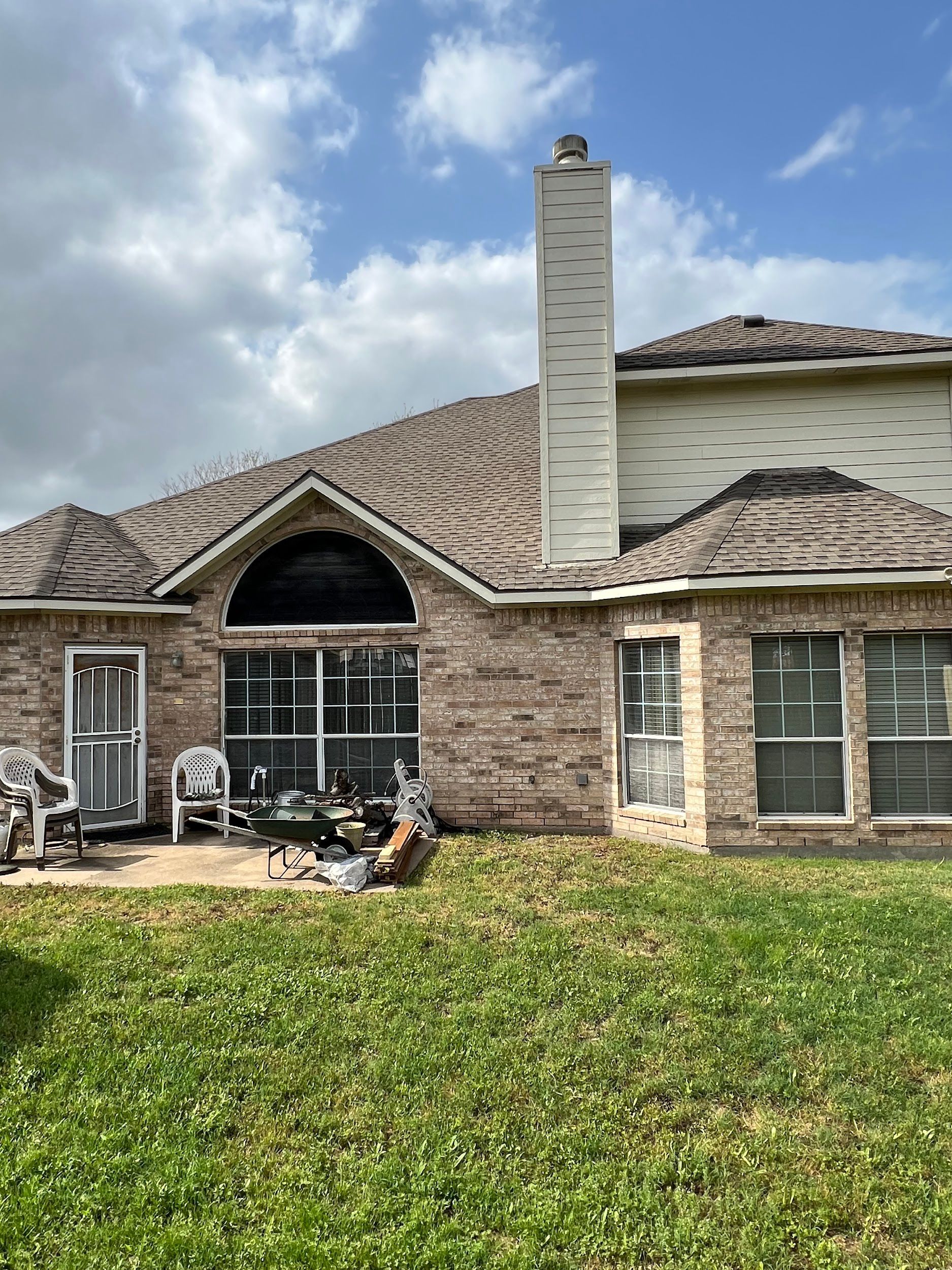 Backyard of a brick house with a brown roof, chimney, and green grass. Patio furniture sits on the patio.
