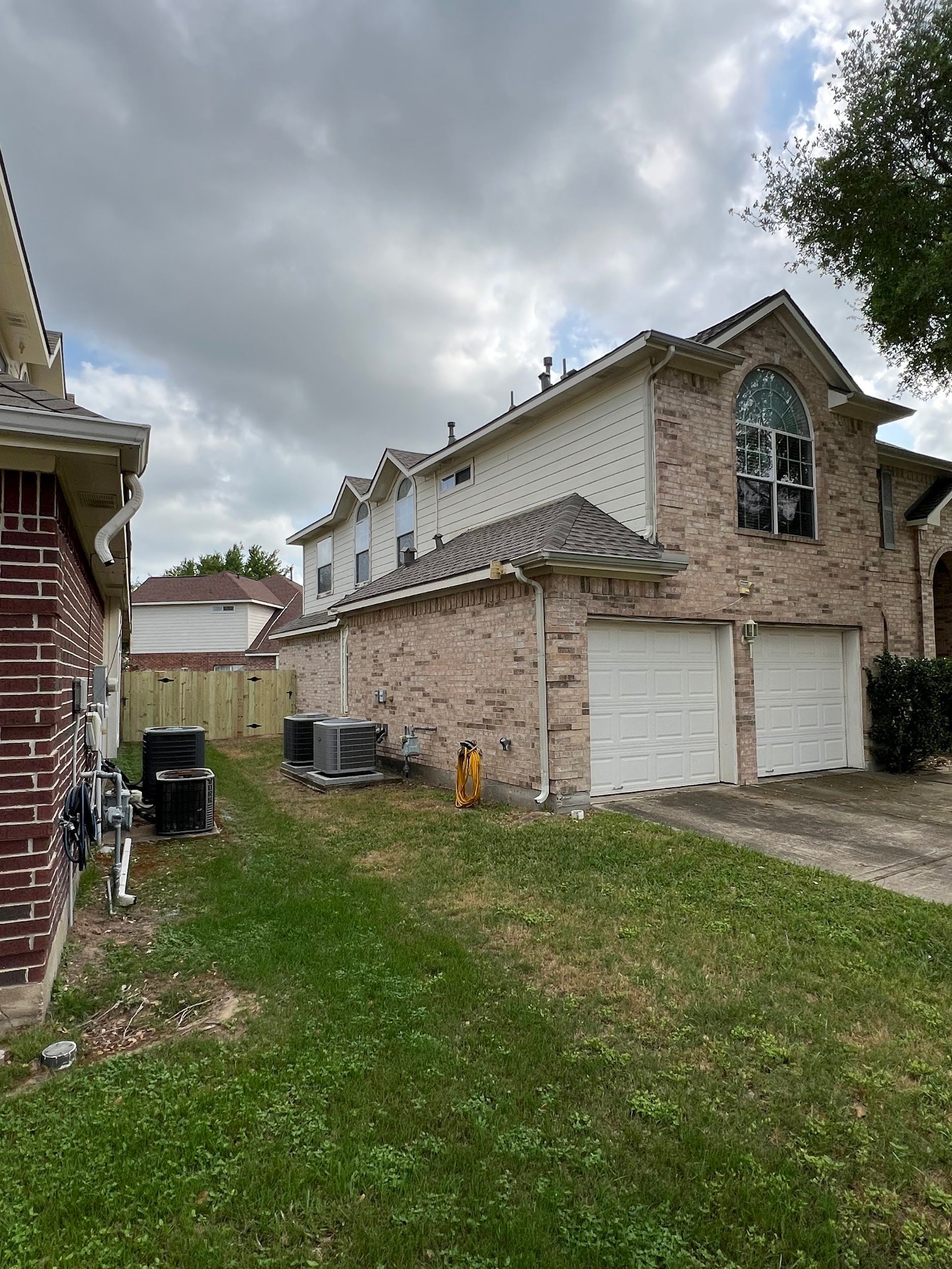 Exterior of a two-story brick house with a two-car garage, grass lawn, and a wooden fence on a cloudy day.