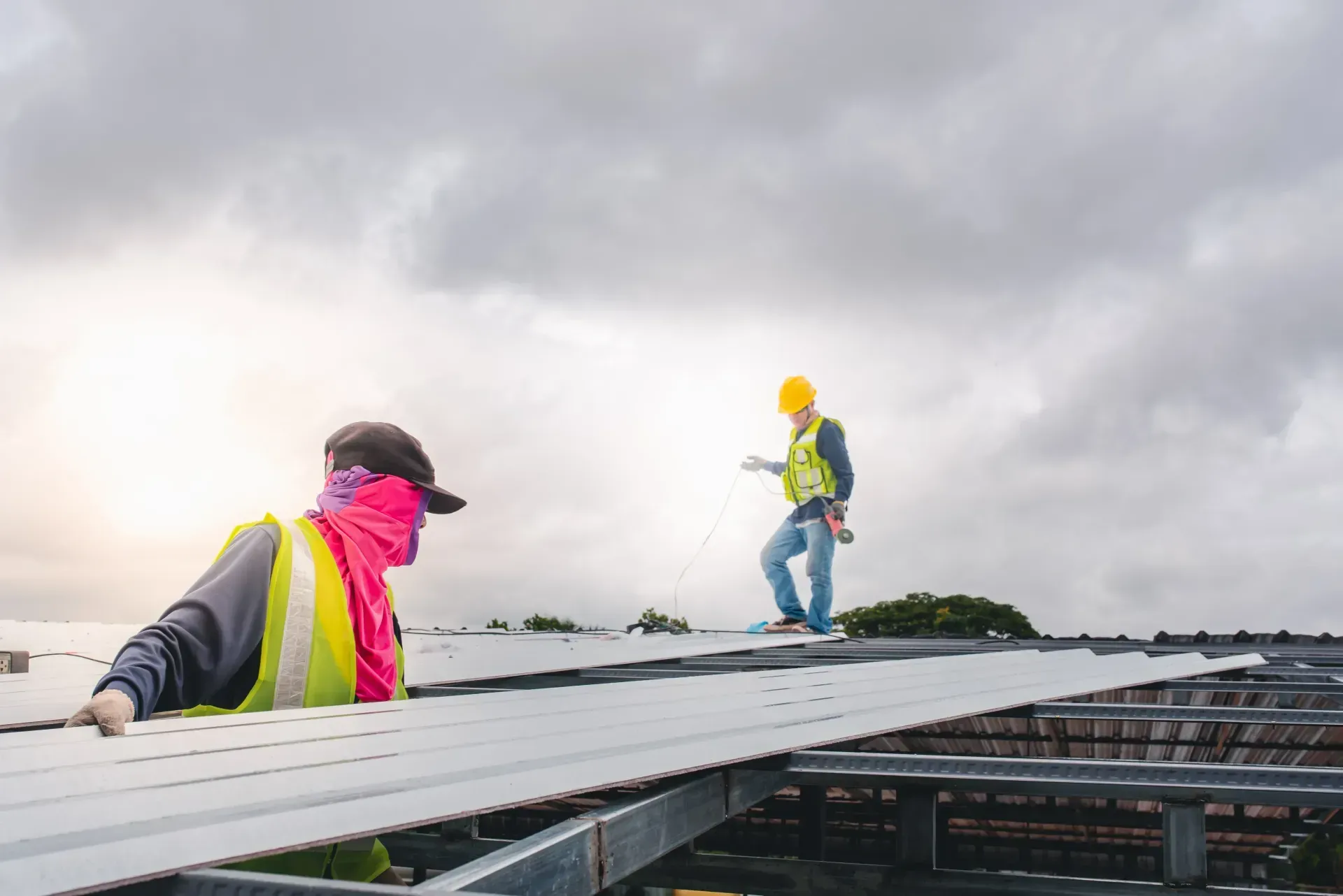 Construction workers installing metal panels on a roof under cloudy sky. One worker wears a yellow vest and hard hat.