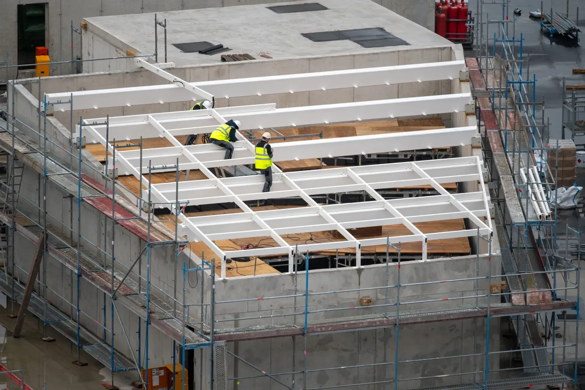 Construction workers on scaffolding, building roof frame. Wearing yellow vests, concrete walls.