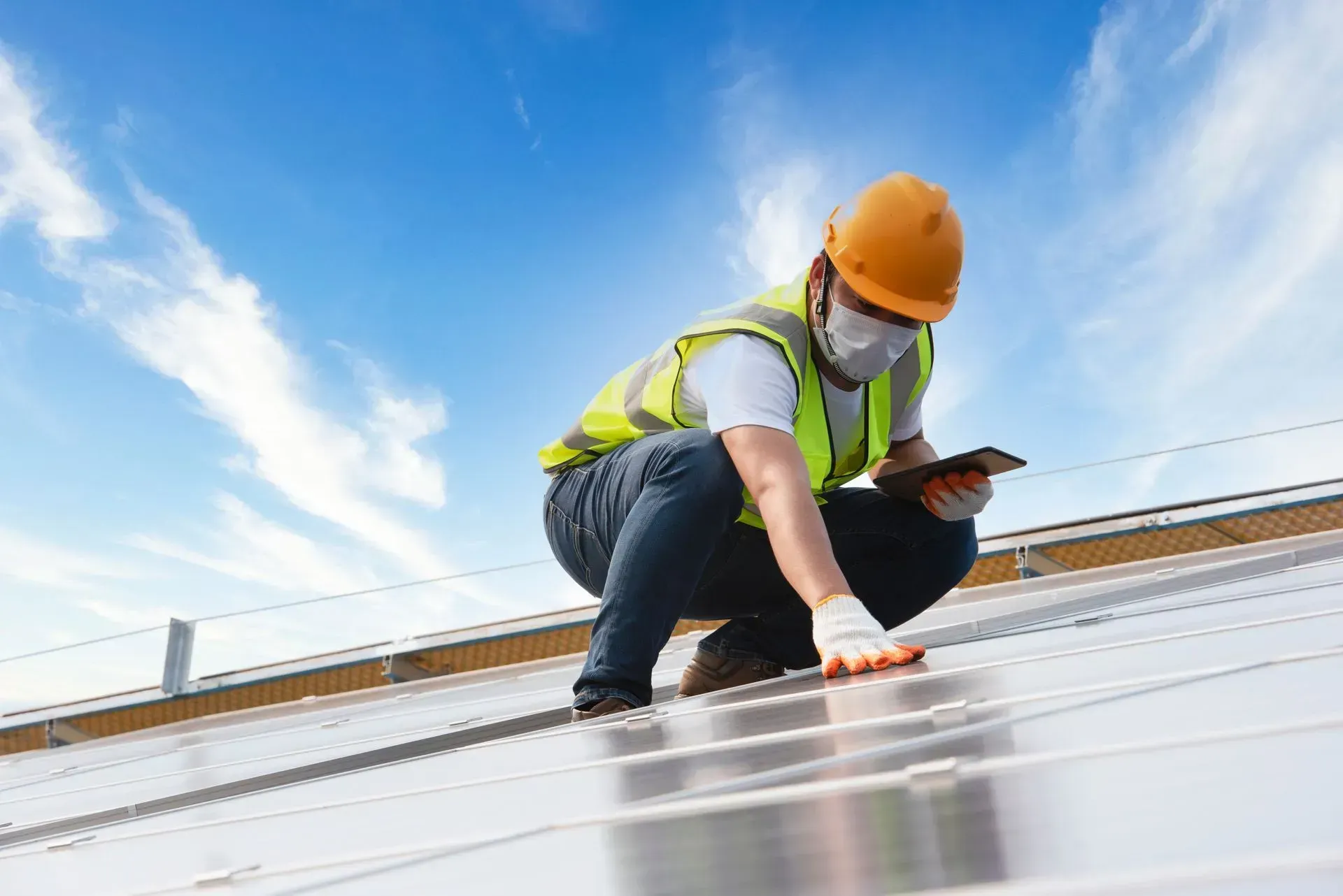Construction worker inspects solar panels on a rooftop, wearing a safety helmet, vest, gloves, and mask under a blue sky.