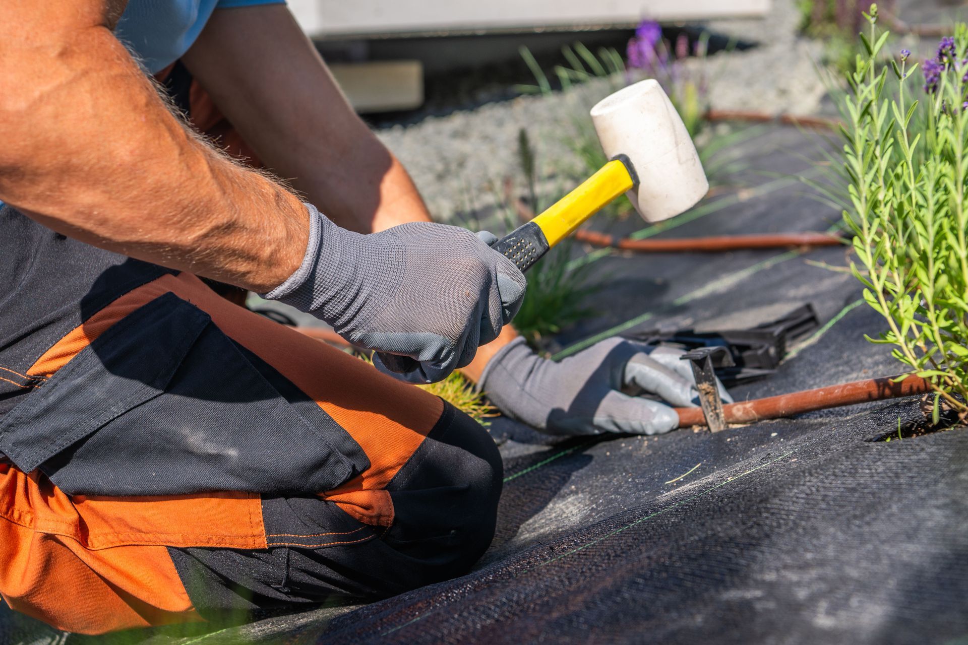 Person using a mallet to secure a tube to landscape fabric in a garden, wearing gloves.
