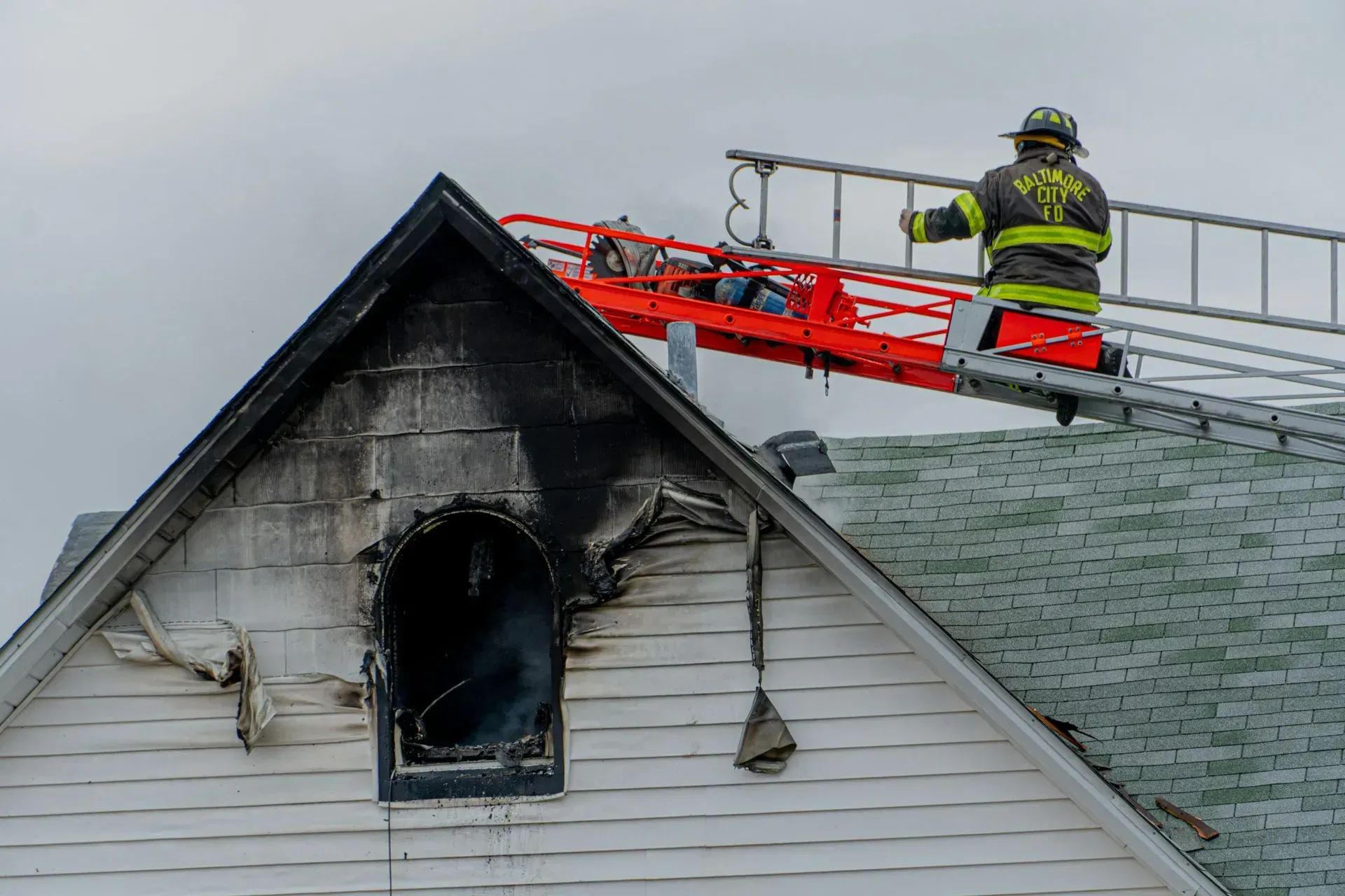 Firefighter on a ladder next to a burning house, with a damaged window and smoke.