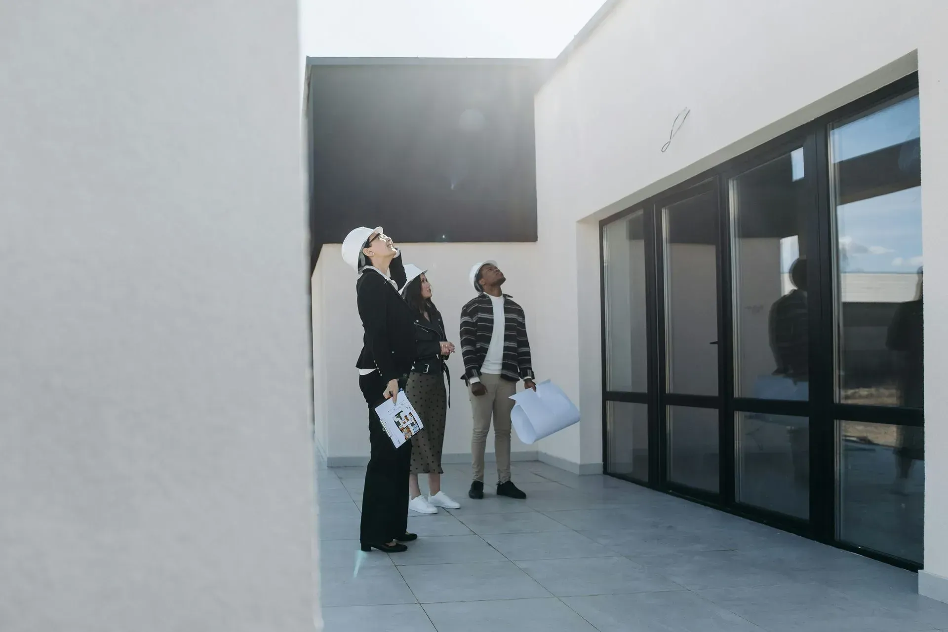 Three people, including one wearing a hard hat, inspecting the interior of a building with large windows.